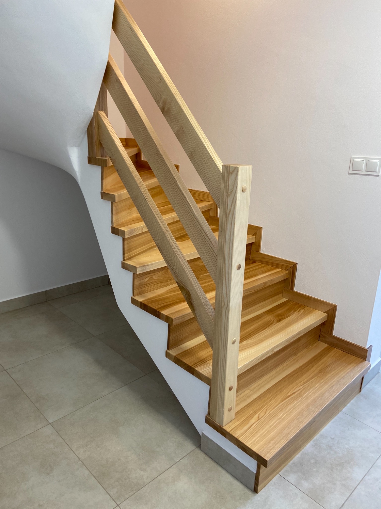 Jasne, drewniane schody z barierką, zrealizowane w Czarna Góra, prowadzące na górę, widoczne na tle białych ścian i płytek podłogowych.