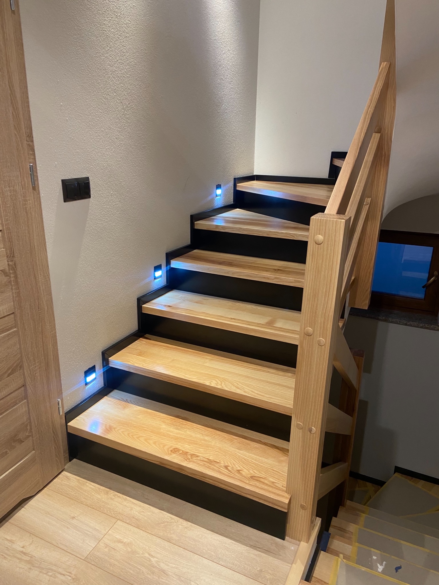 Drewniane schody z podświetleniem LED w ścianie, czarne podstopnice, jasne drewno, widoczna balustrada i fragment drzwi.