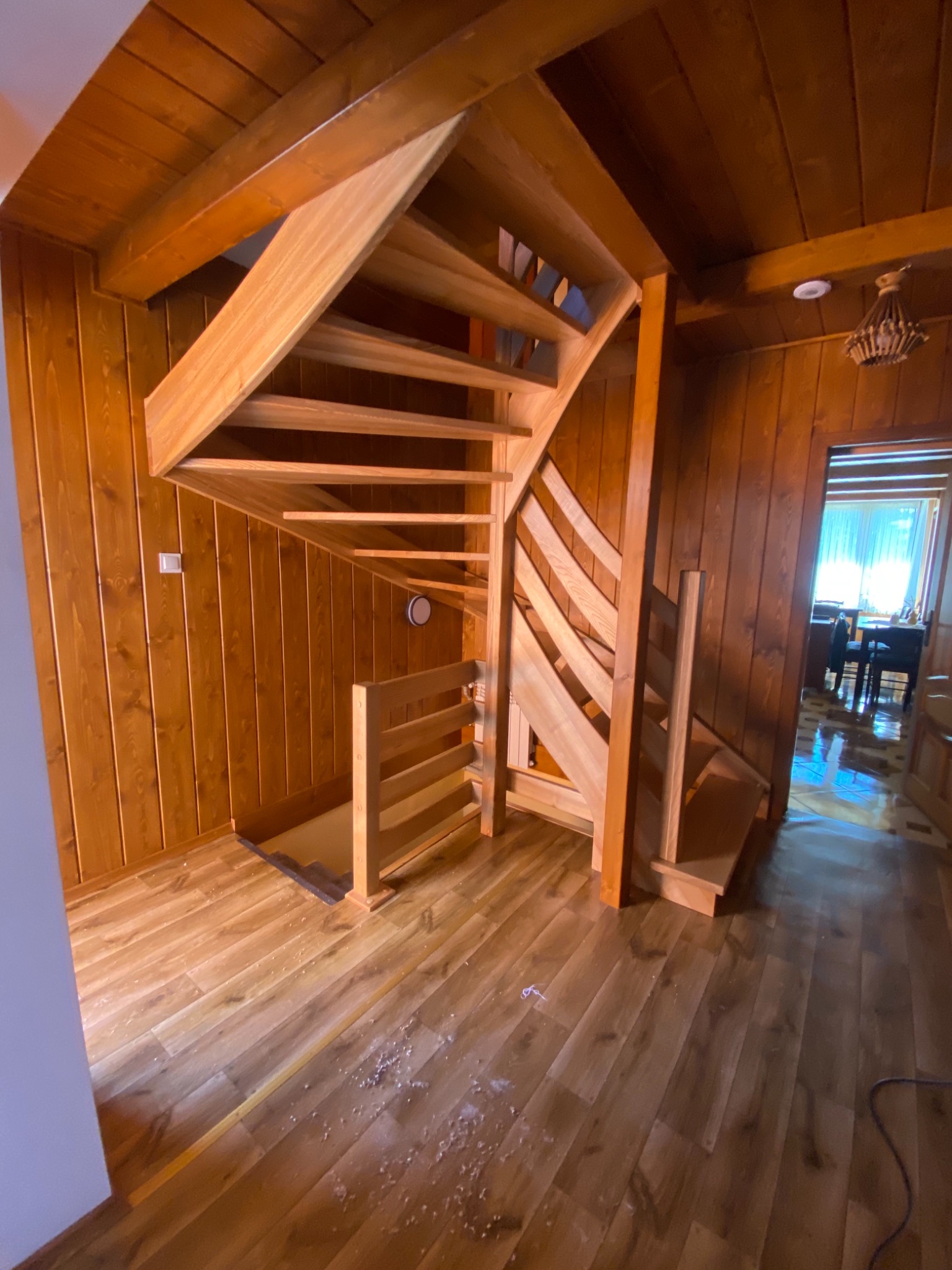 Spiralne, drewniane schody z jasnego drewna w pomieszczeniu wykończonym boazerią, widoczne wióry na podłodze.