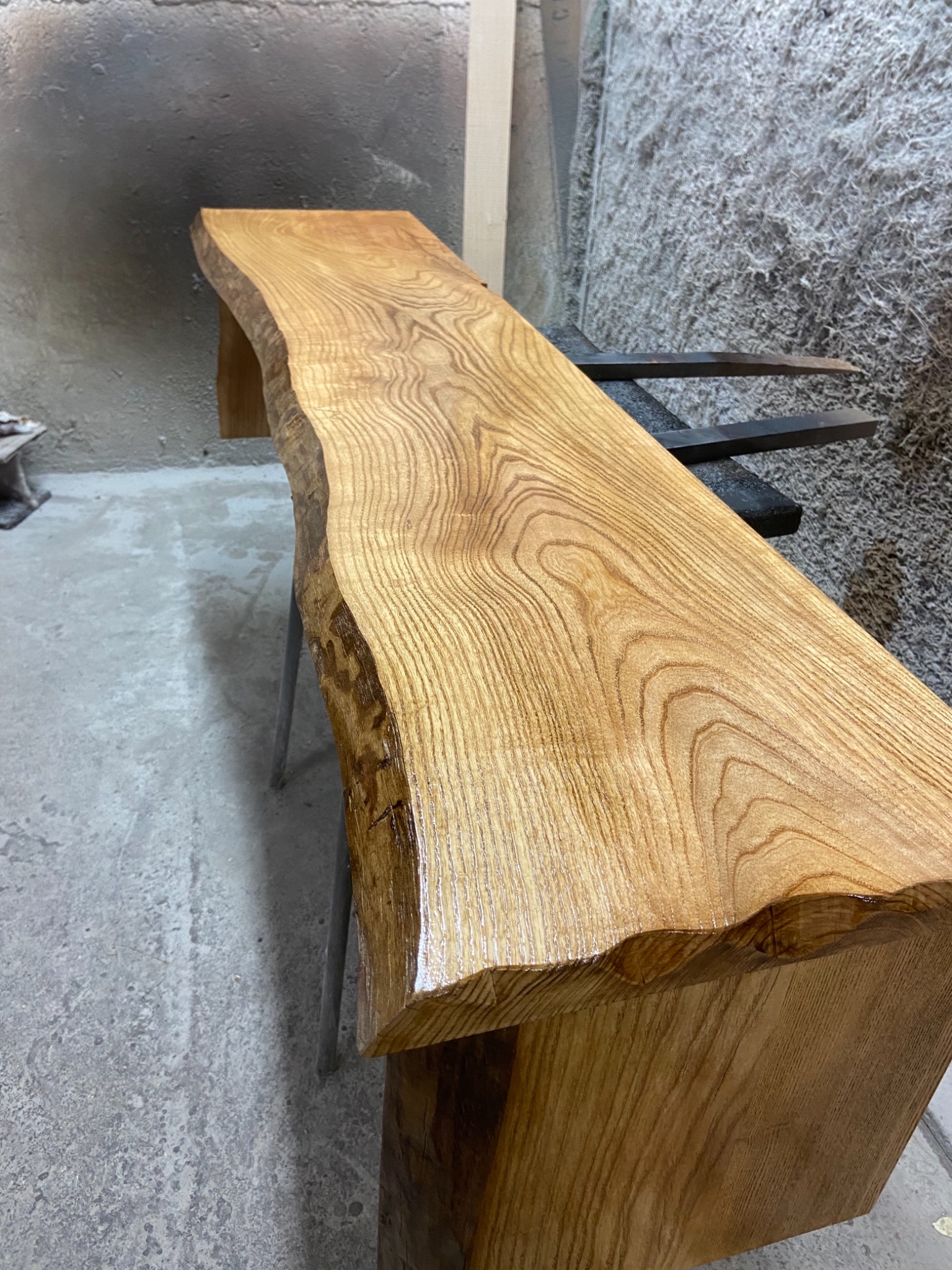 Surowy, lakierowany blat z litego drewna o nieregularnych krawędziach, ustawiony na metalowych nogach w warsztacie.