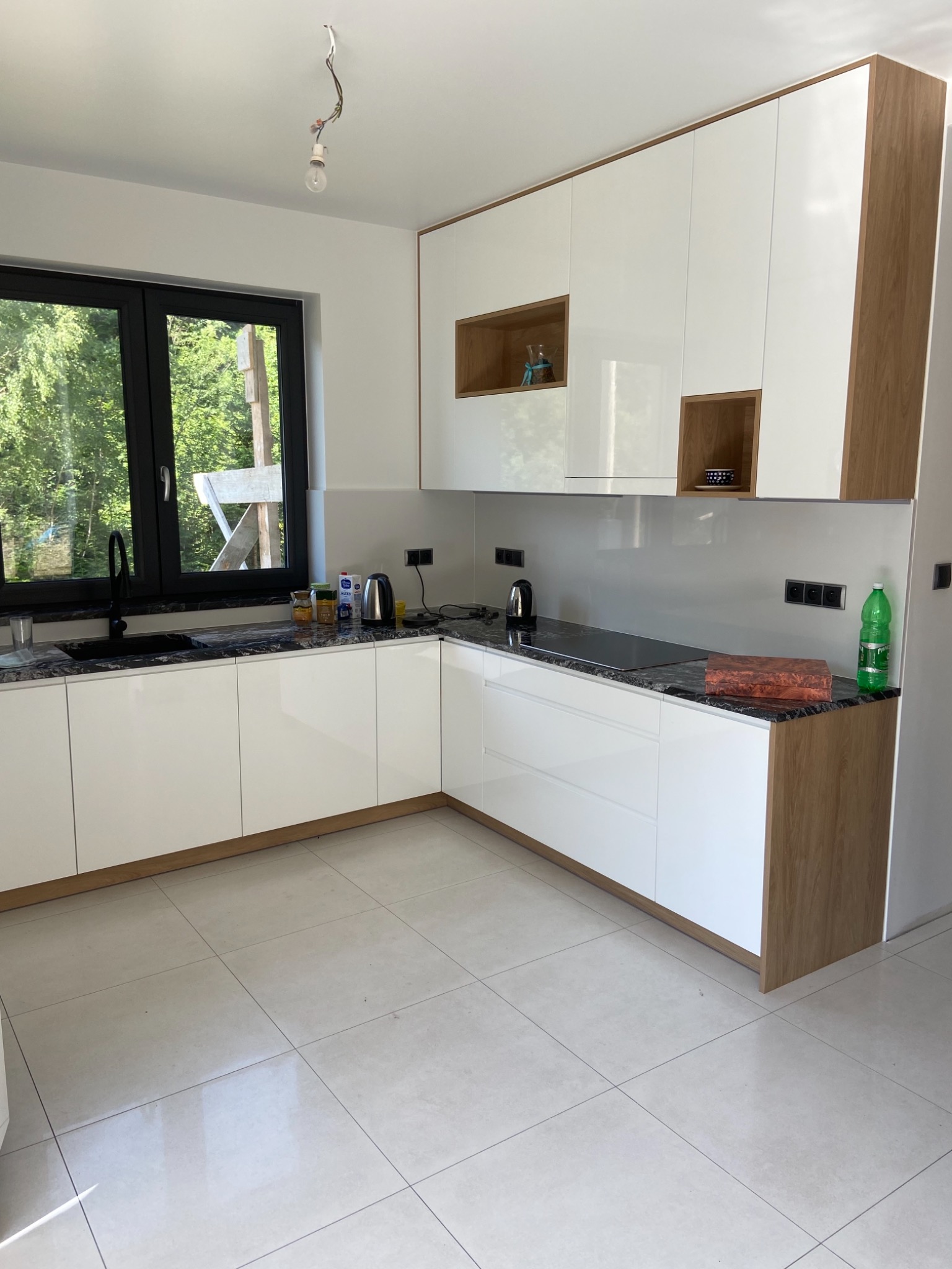 Nowoczesna kuchnia z białymi frontami i drewnianymi akcentami, czarnym blatem z kamienia, czarnym oknem z widokiem na zieleń, wisząca żarówka bez oprawy.