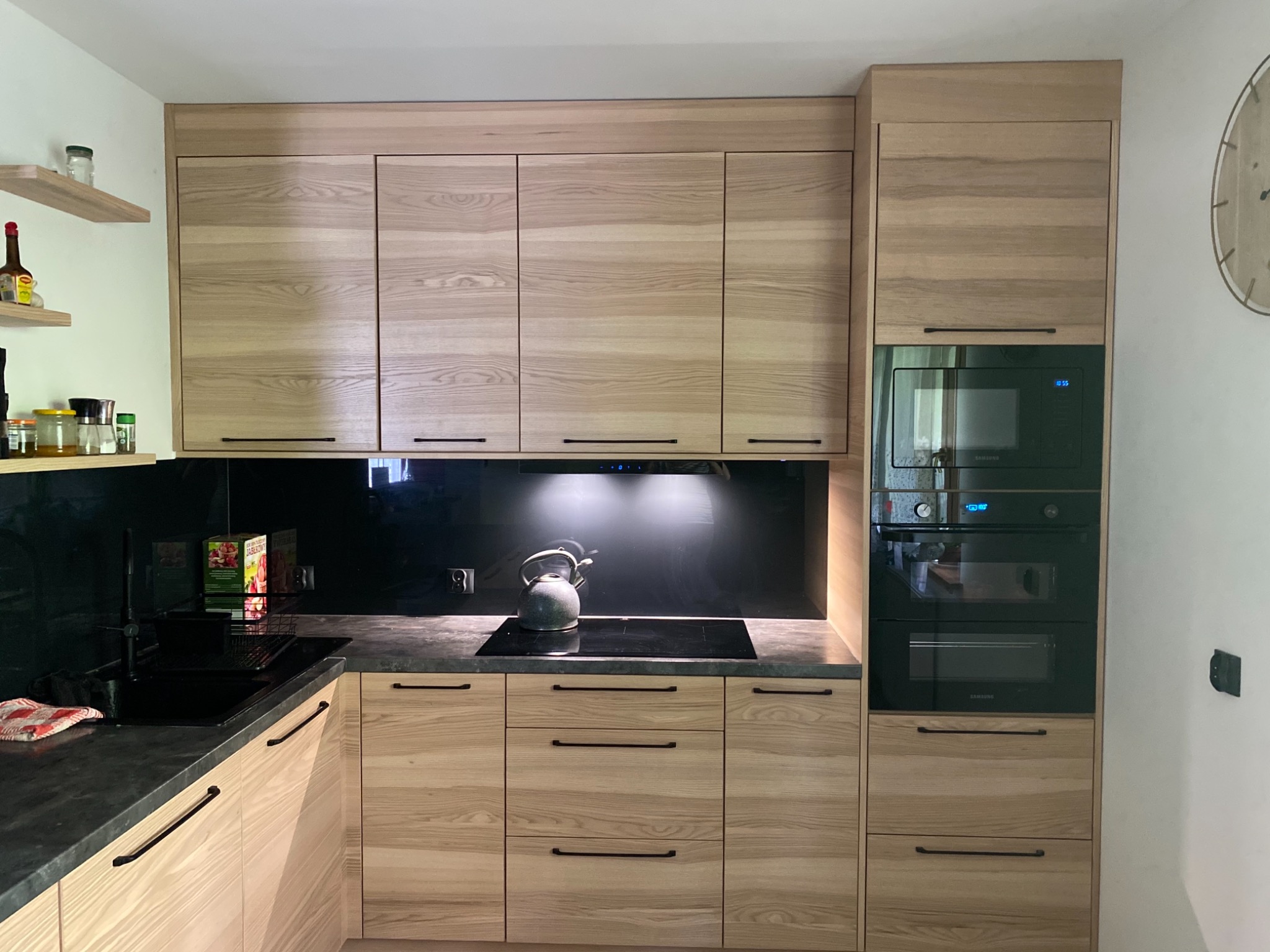 Jasna, nowoczesna zabudowa kuchenna z drewnianymi frontami, czarnym blatem i sprzętem AGD w zabudowie, widok na płytę indukcyjną z czajnikiem.