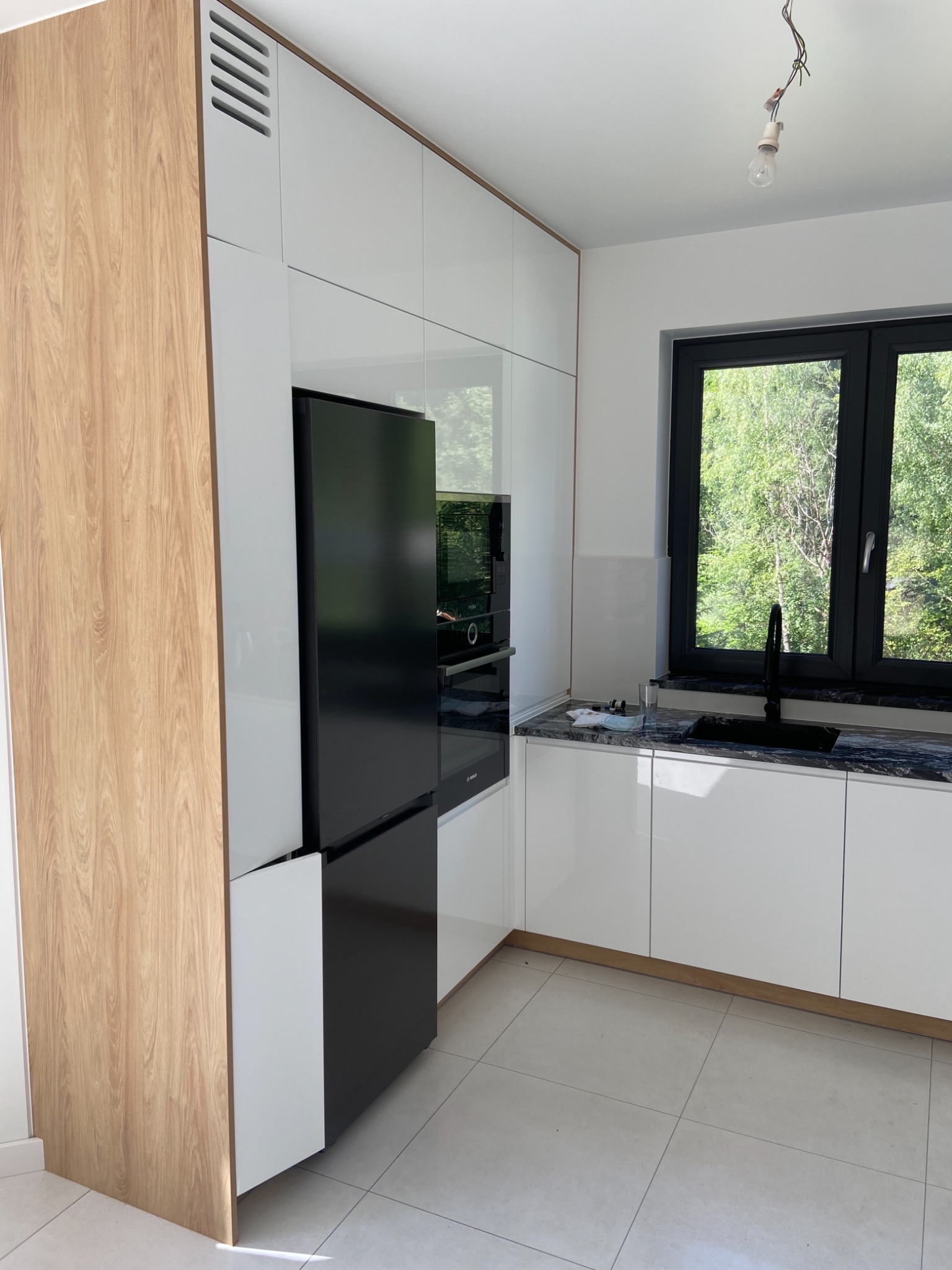 Nowoczesna kuchnia z białymi szafkami, czarnym zlewem i armaturą, drewnianym wykończeniem i czarną lodówką w zabudowie, widok na zieleń za oknem, niedokończone oświetlenie sufitowe.