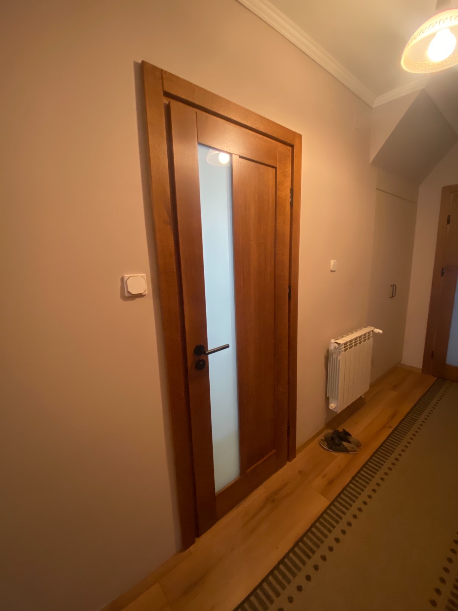 Brązowe drzwi wewnętrzne z szybą z matowego szkła, zamontowane w korytarzu z drewnianą podłogą i beżowymi ścianami, widoczny grzejnik i pantofle.