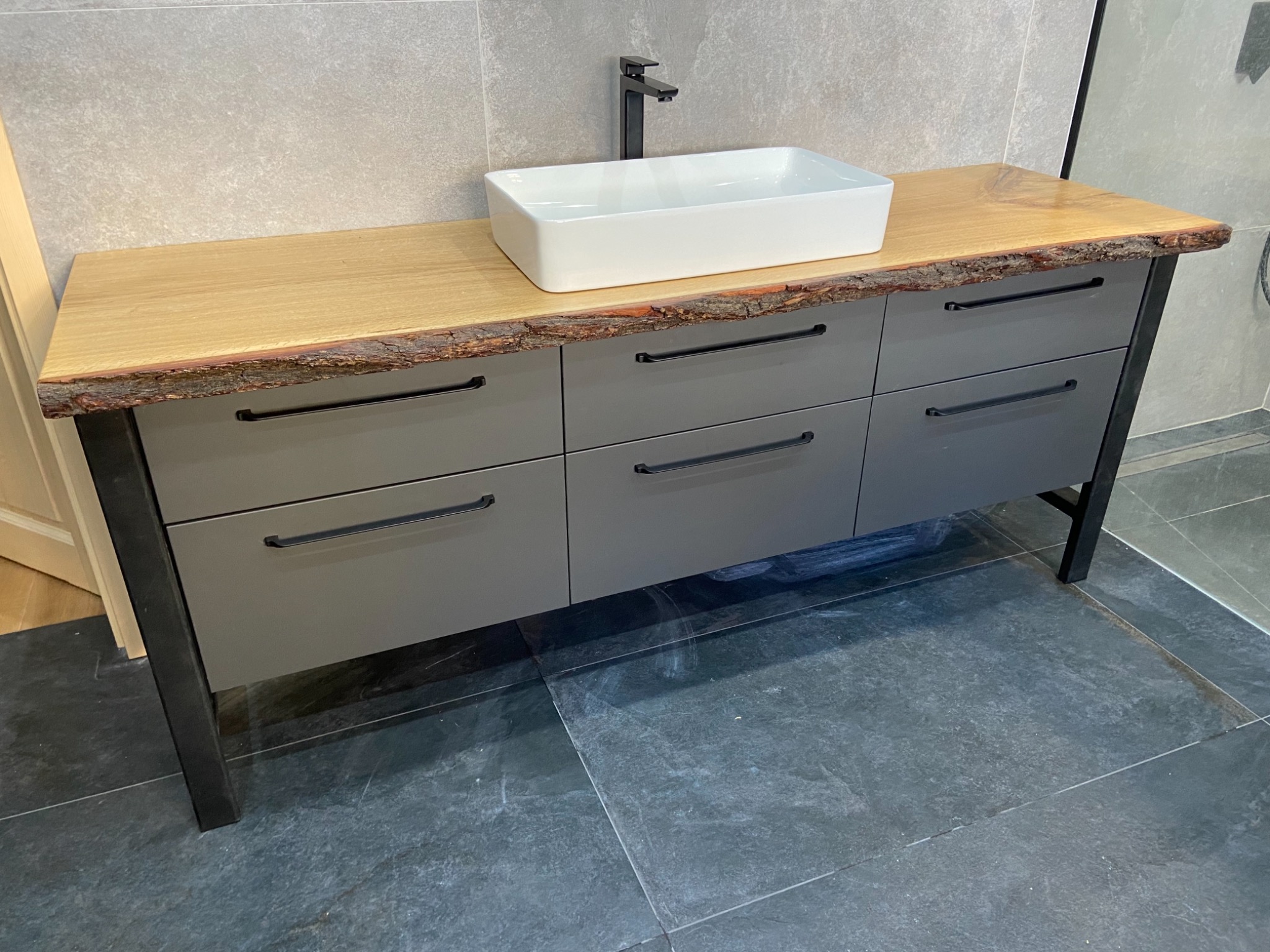 Szara szafka łazienkowa z sześcioma szufladami, blatem z naturalnego drewna z korą i białą umywalką nablatową, czarna armatura i grafitowa podłoga.