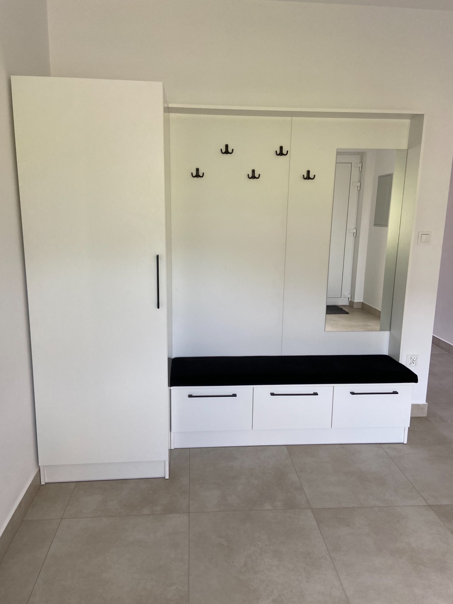 Biała zabudowa przedpokoju z szafą, siedziskiem z czarną poduszką i lustrem, minimalistyczne wieszaki na ścianie.