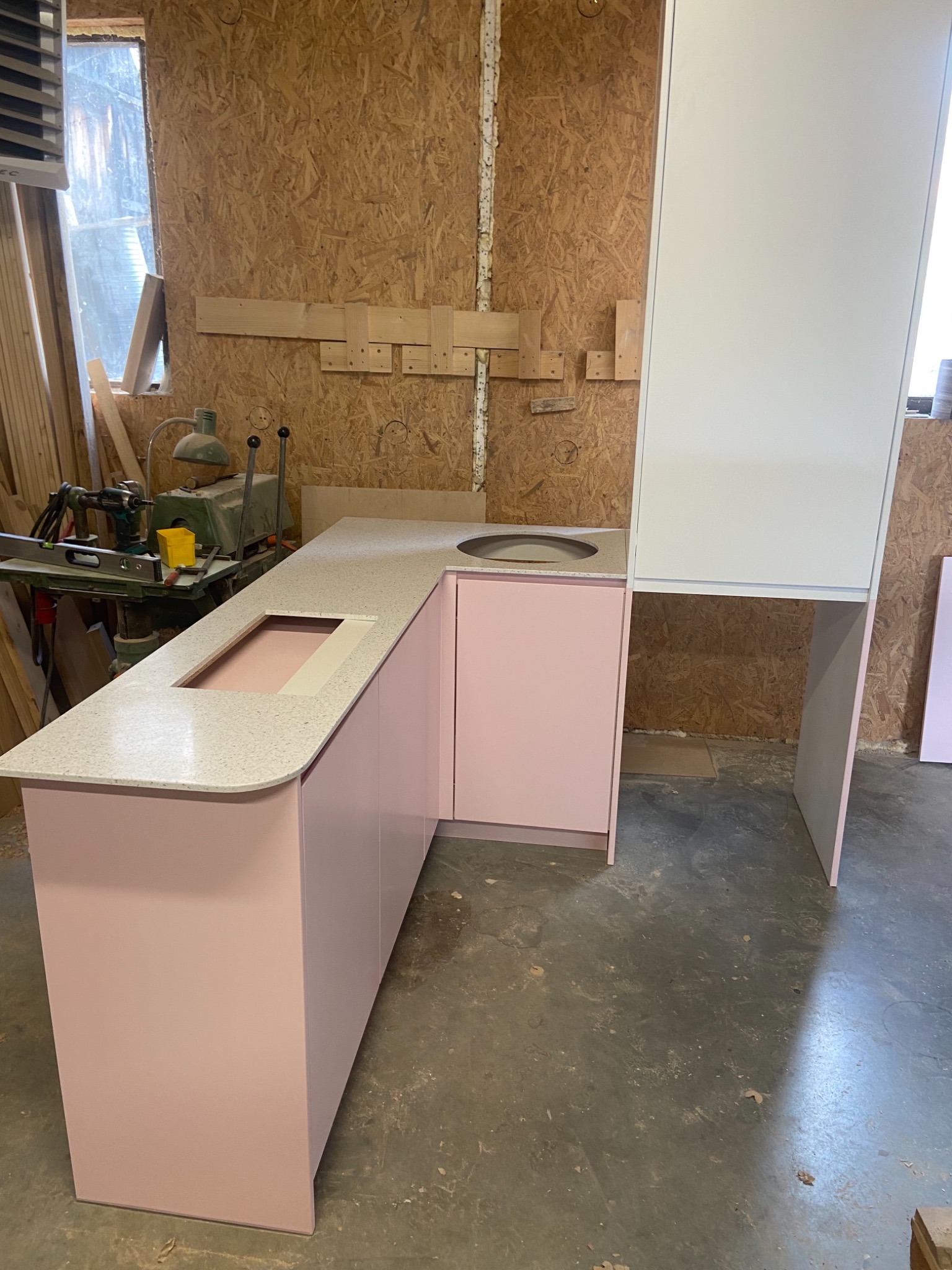 Różowa zabudowa kuchenna z blatem z jasnego kamienia w warsztacie, widoczna maszyna stolarska w tle
