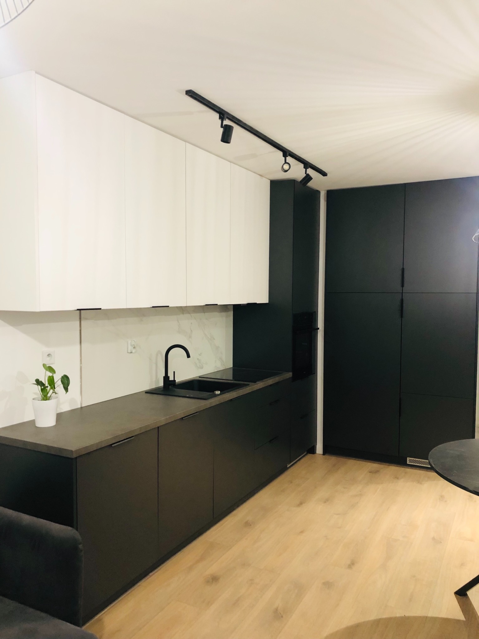 Nowoczesna kuchnia z białymi szafkami górnymi i ciemnoszarymi dolnymi, czarna armatura i oświetlenie punktowe, marmurowy backsplash, roślina doniczkowa na blacie.