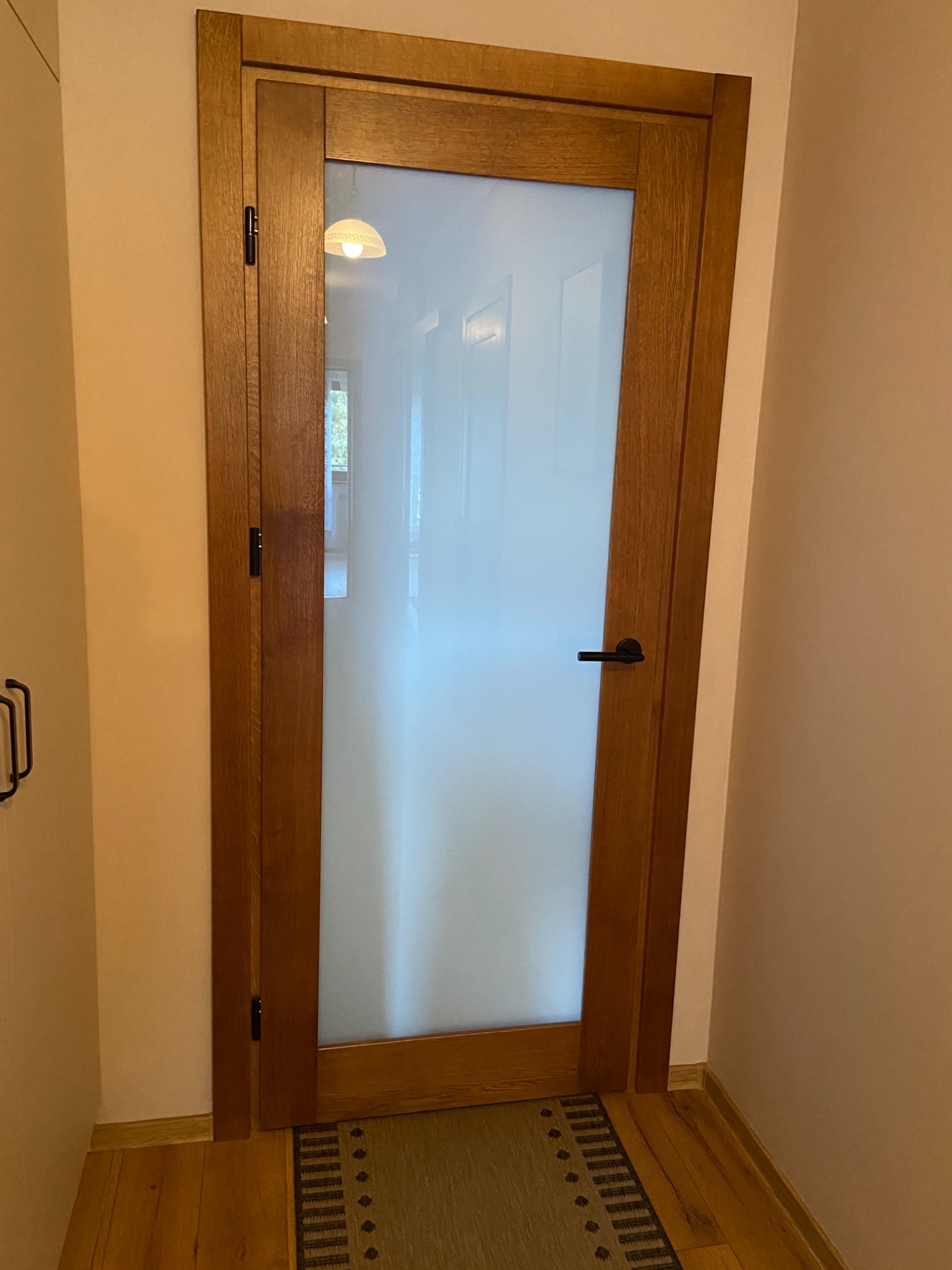 Drewniane drzwi wewnętrzne z matową szybą, czarna klamka i widoczny fragment podłogi z dywanikiem.