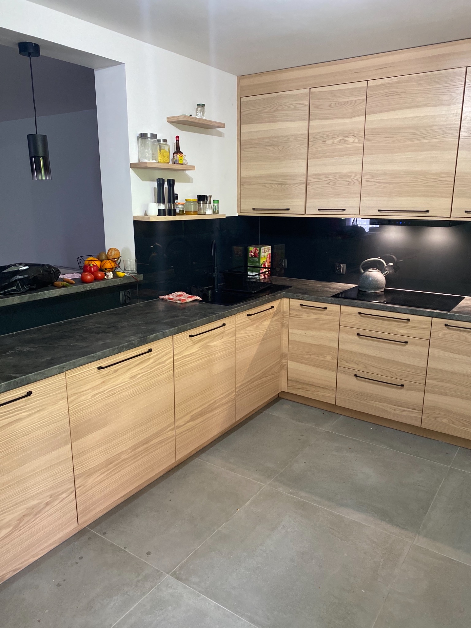 Jasne drewniane szafki kuchenne z czarnym blatem i backsplashiem, minimalistyczne uchwyty, widok na narożnik kuchni z elementami dekoracyjnymi na półkach.