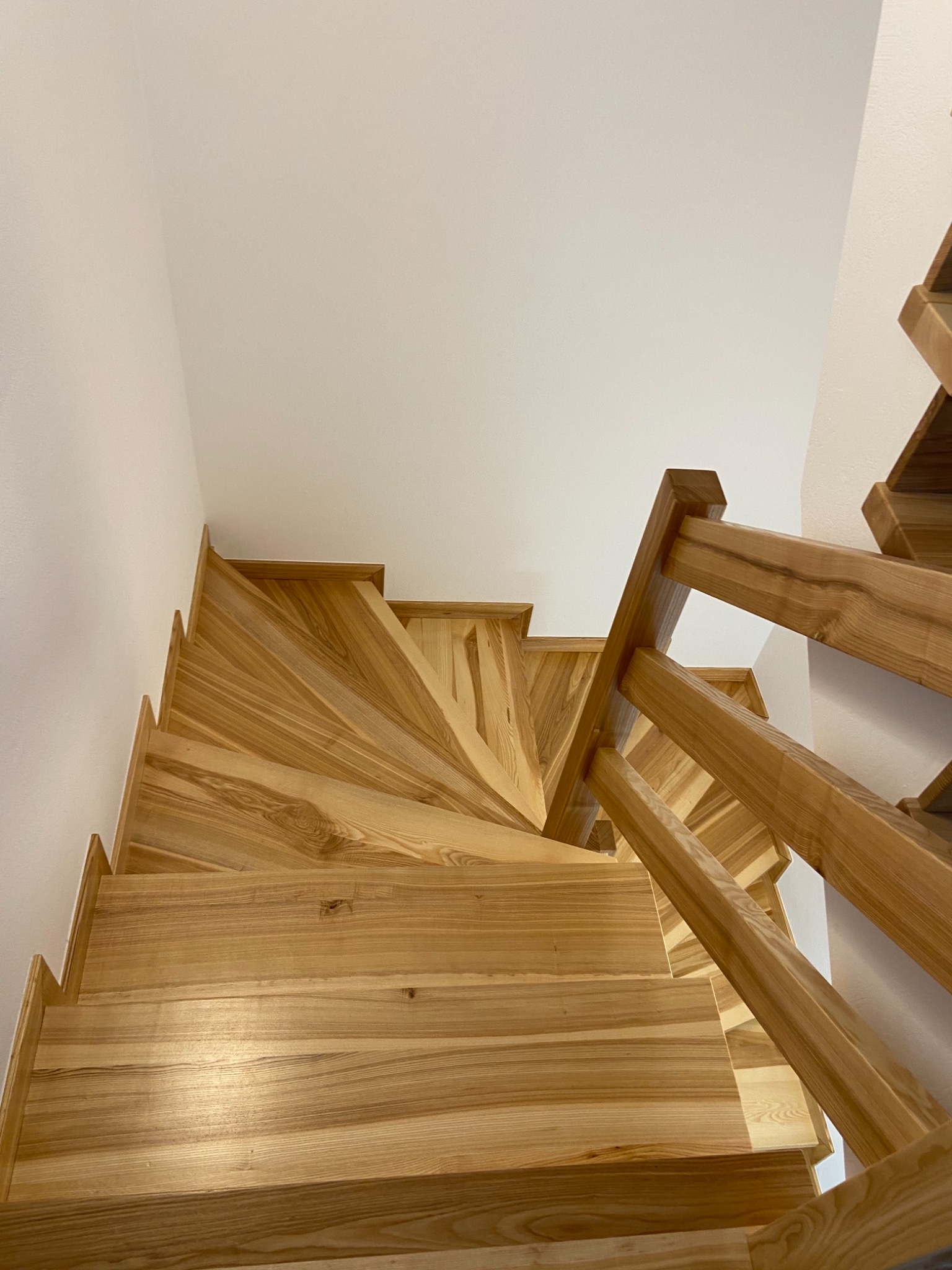 Kątowe ujęcie spiralnych schodów z jasnego drewna z widocznym rysunkiem słojów, zamontowanych w białym wnętrzu.