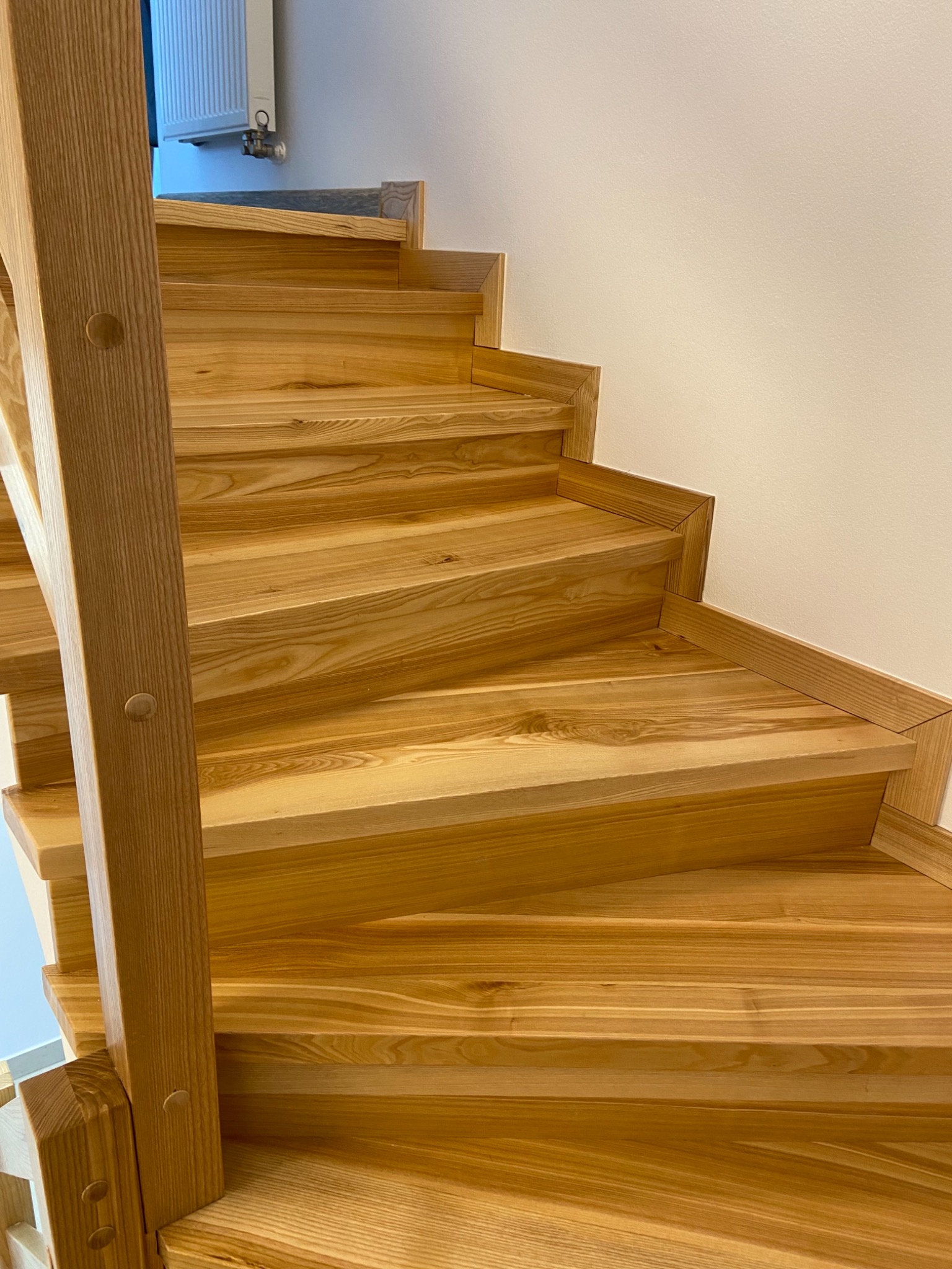 Kątowe ujęcie na jasne, drewniane schody z widoczną strukturą drewna, prowadzące w górę wzdłuż białej ściany, z widocznym fragmentem grzejnika w tle.