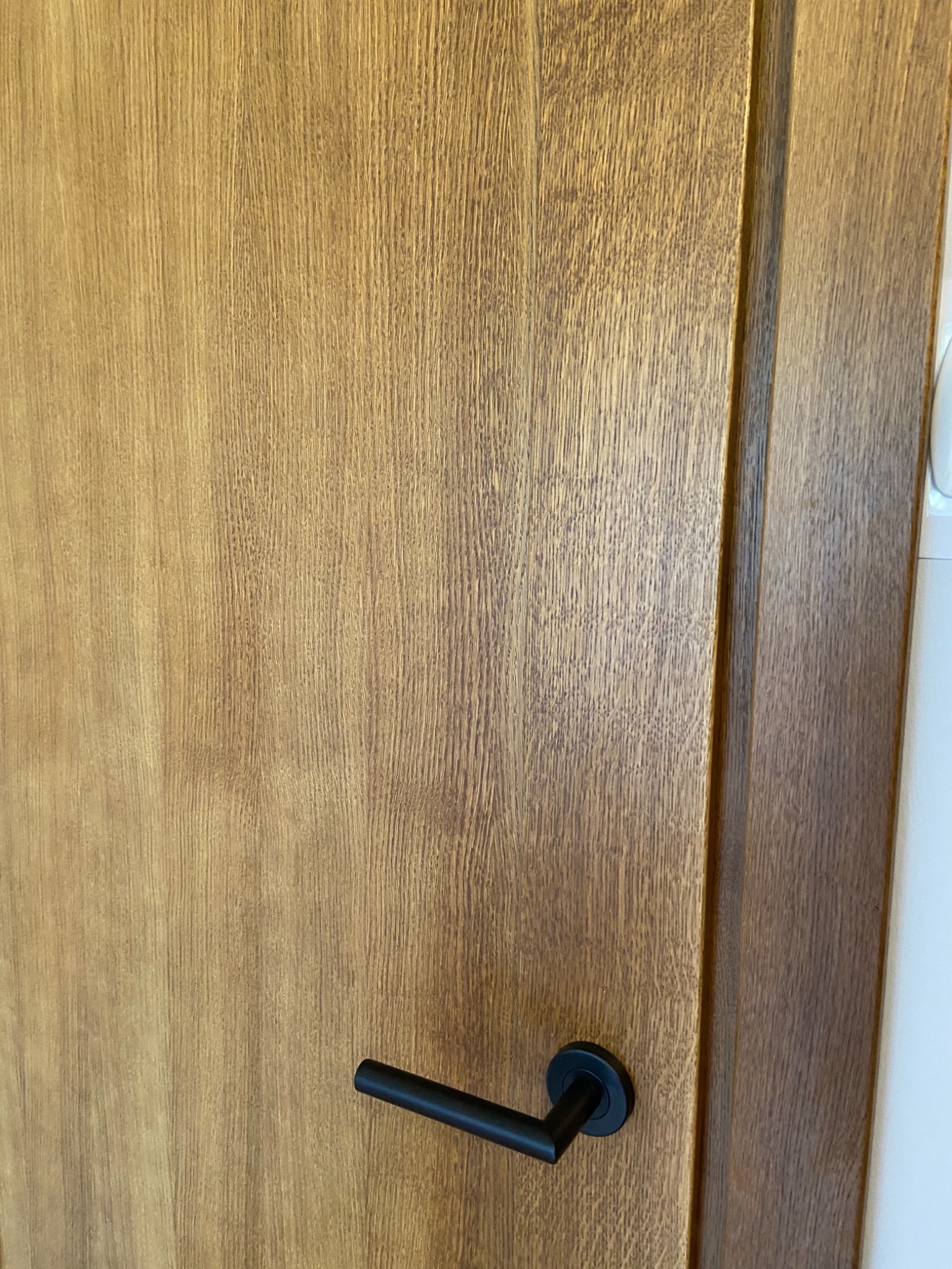 Drewniane drzwi wewnętrzne z czarną, minimalistyczną klamką, widoczna struktura drewna i ościeżnica.