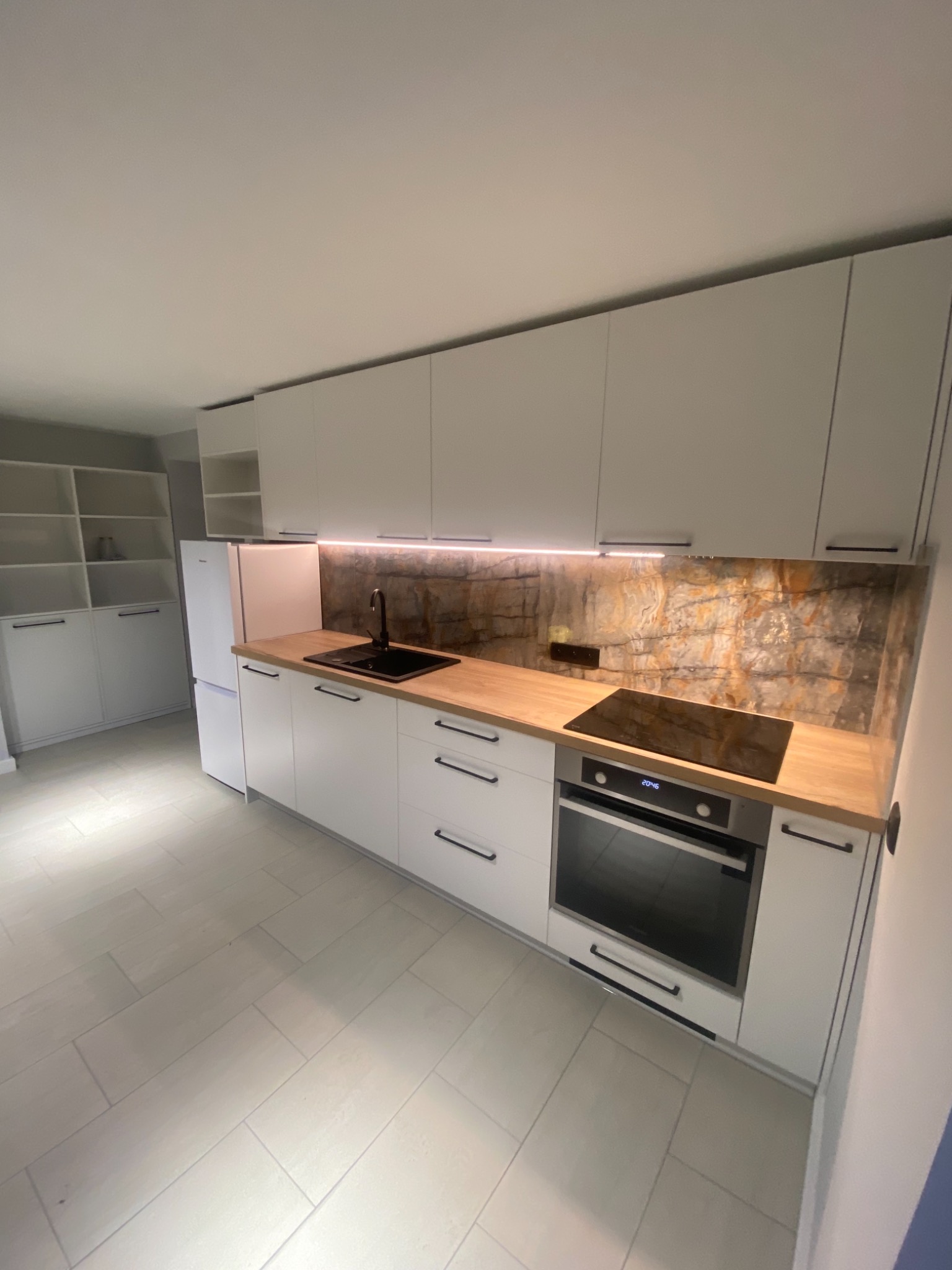 Nowoczesna kuchnia z białymi frontami szafek, drewnianym blatem i kamiennym panelem nad blatem, uzupełniona czarną armaturą i sprzętem AGD.