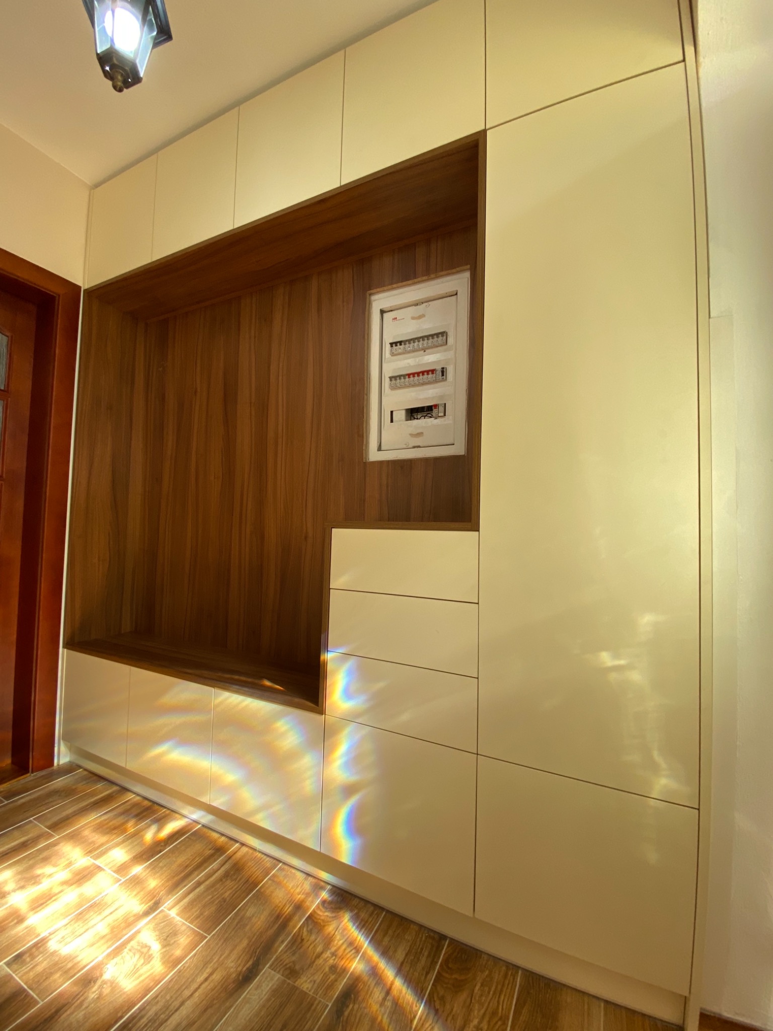 Szafa wnękowa z jasnymi frontami, drewnianym wykończeniem wnęki i siedziskiem, oświetlona światłem słonecznym tworzącym tęczowe refleksy na podłodze i meblach, obok drzwi.