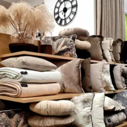 Dekoracyjne poduszki i koce w odcieniach beżu i brązu ułożone na drewnianych półkach, w tle zegar i zasłony