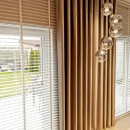 Beżowe zasłony z aksamitnego materiału, wiszące obok białych żaluzji drewnianych, oświetlone nowoczesną lampą z kulistymi kloszami.