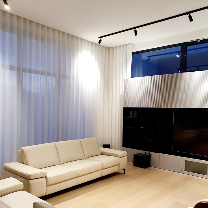 Jasny salon z beżową sofą, białymi, transparentnymi zasłonami i nowoczesną zabudową ściany z telewizorem, oświetlony reflektorami sufitowymi.