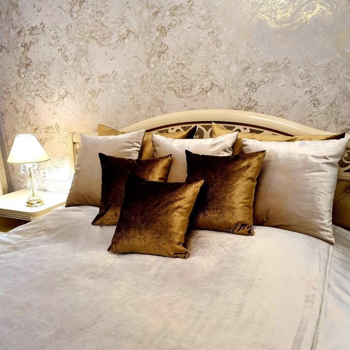 Eleganckie łóżko z kremową pościelą i dekoracyjnymi poduszkami w odcieniach beżu i brązu, sfotografowane w Koszalinie. Na stoliku nocnym kryształowa lampka.