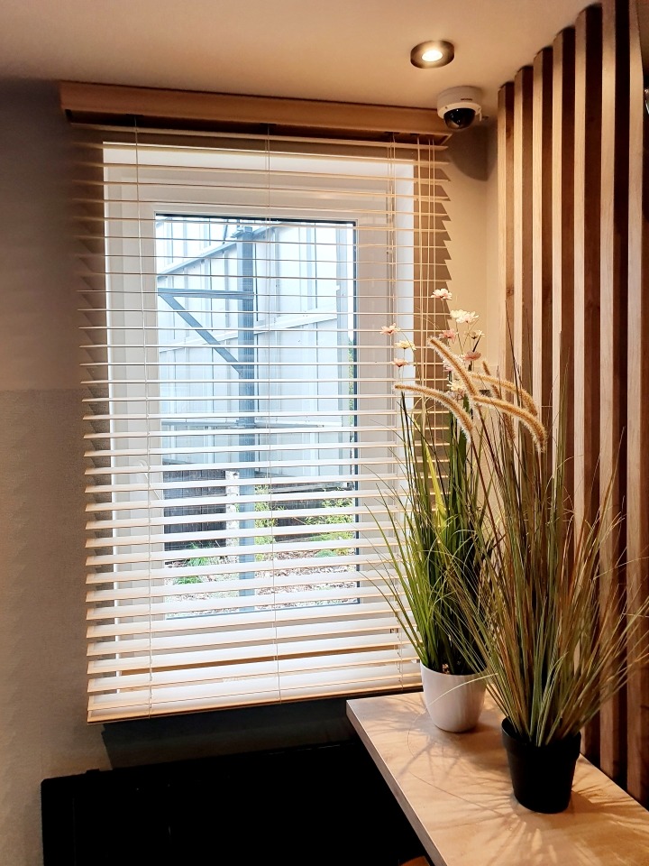 Drewniane żaluzje poziome w jasnym oknie z widokiem na konstrukcję szklarni; dekoracyjne trawy w doniczkach na blacie, drewniana okładzina ścienna.