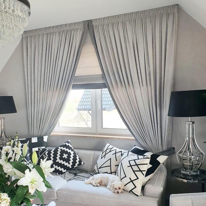 Jasnoszara roleta rzymska i zasłony z ciężkiej tkaniny w oknie na poddaszu, dekoracyjne poduszki na sofie i mały pies rasy Maltańczyk.