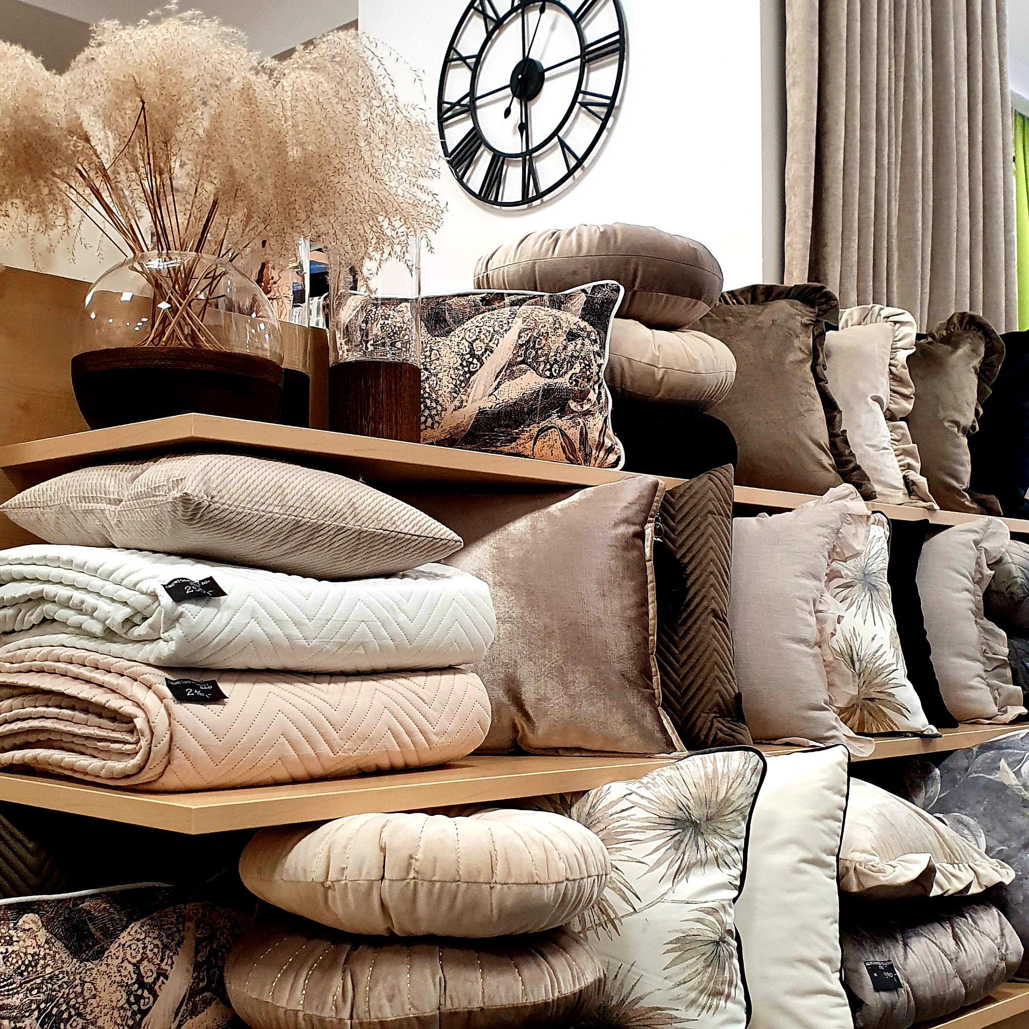 Dekoracyjne poduszki i koce w odcieniach beżu i brązu ułożone na drewnianych półkach, w tle zegar i zasłony