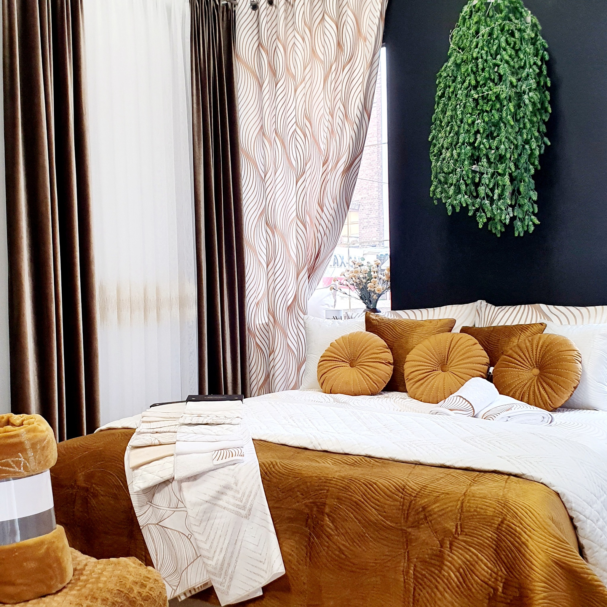 Elegancka sypialnia z brązowymi i białymi zasłonami w geometryczne wzory, okrągłymi poduszkami w kolorze musztardowym oraz dekoracyjną rośliną pnącą na czarnej ścianie.