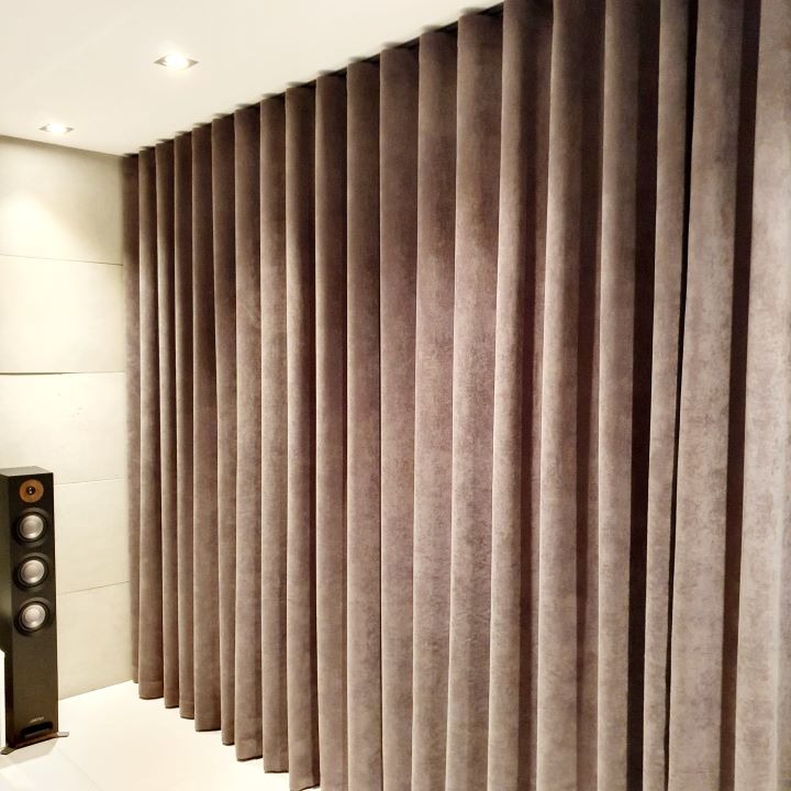 Długie, marszczone zasłony w odcieniu taupe, wiszące od sufitu do podłogi, z widocznym głośnikiem kolumnowym po lewej stronie i minimalistycznym oświetleniem sufitowym.