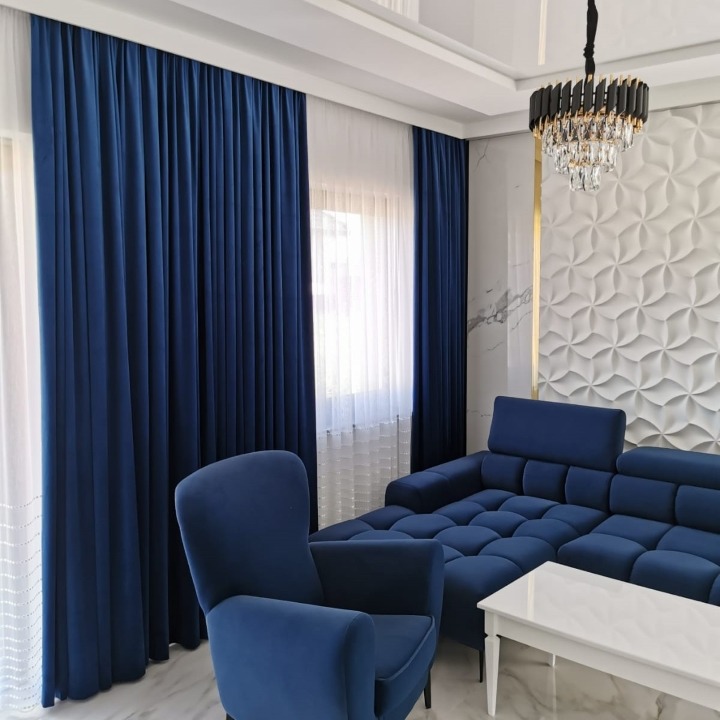 Eleganckie, granatowe zasłony z grubego materiału, zestawione z białymi, zwiewnymi firanami, dopełniają aranżację nowoczesnego salonu z teksturowaną ścianą i kryształowym żyrandolem.