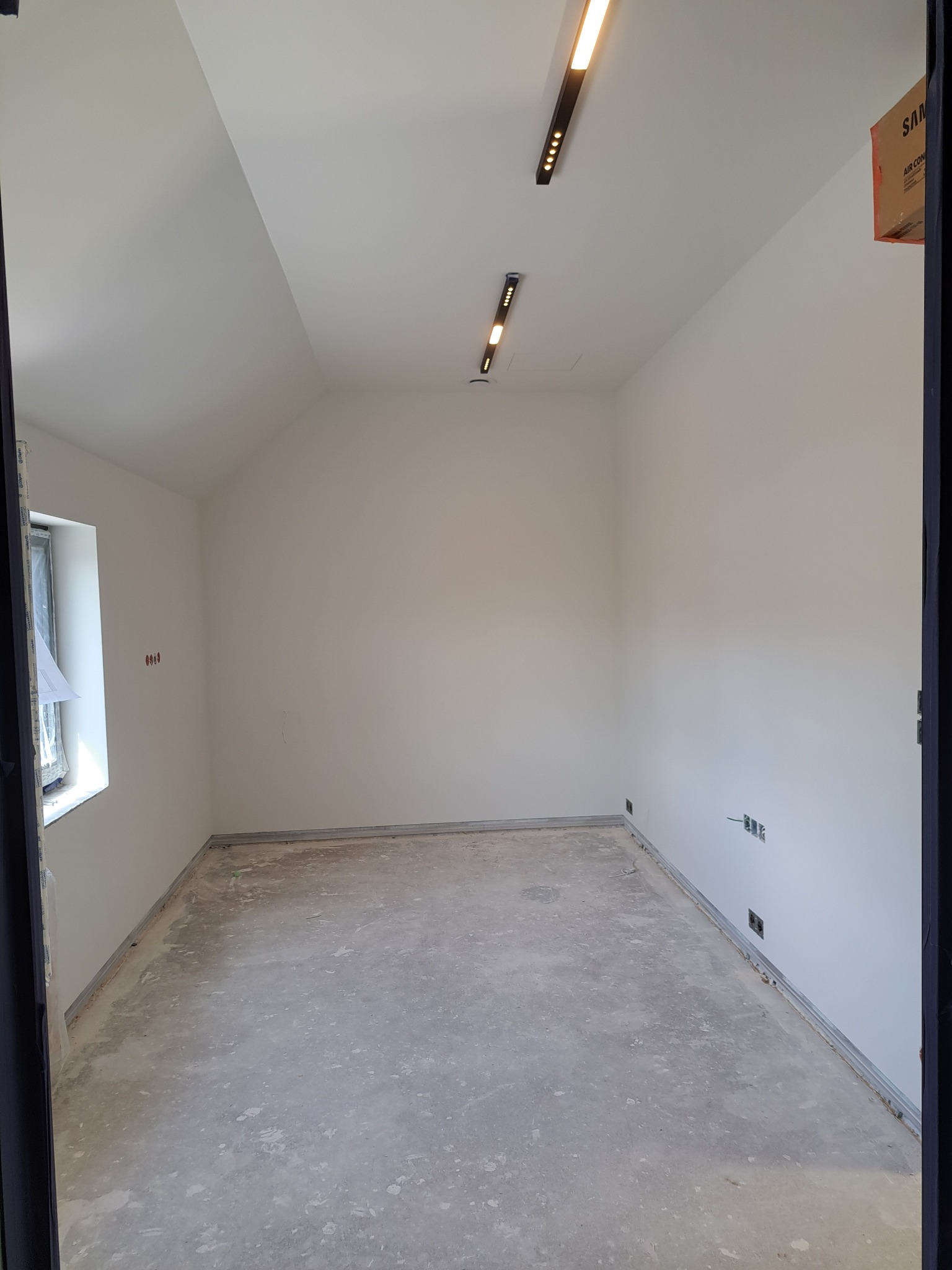 Pusty pokój z białymi ścianami i betonową podłogą przygotowany do dalszych prac wykończeniowych, widoczne listwy przypodłogowe i oświetlenie sufitowe