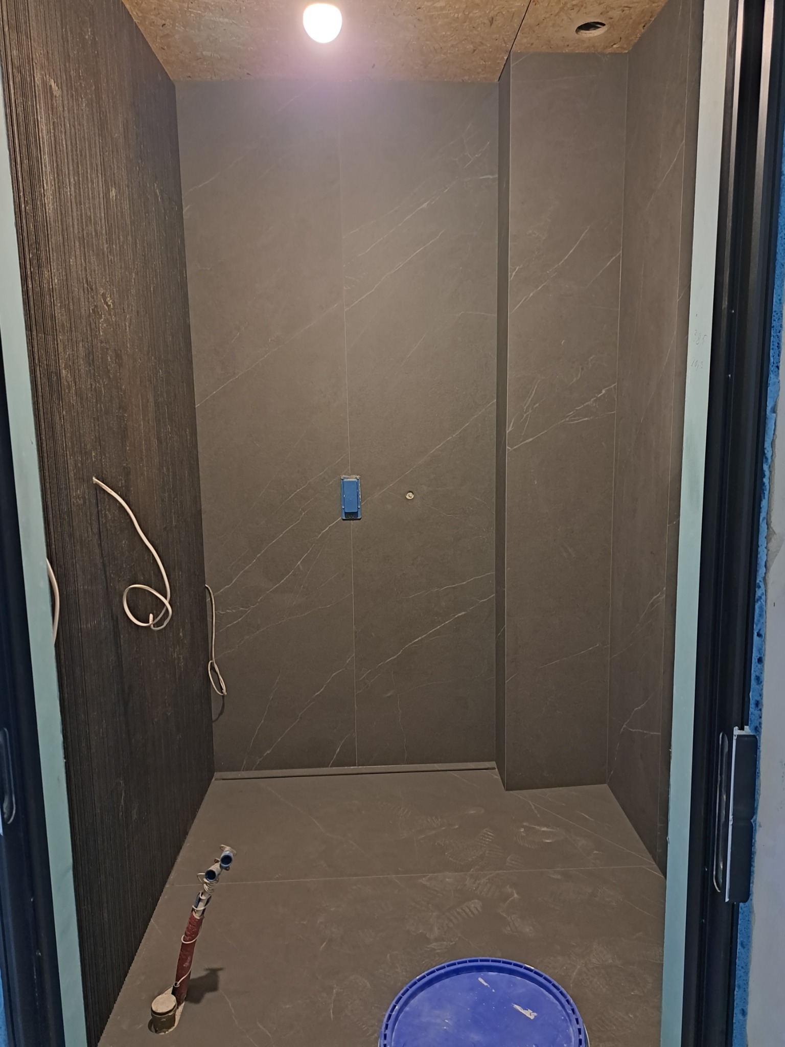 Wnętrze kabiny prysznicowej w trakcie wykańczania, z ciemnymi płytkami imitującymi kamień, widoczne wystające kable i rury, niebieskie wiadro na podłodze i surowy sufit z płyty OSB.