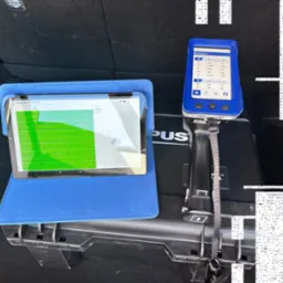 Tablet z otwartą aplikacją prezentującą listę katalizatorów z cenami, obok analizator metali z wynikami pomiarów w bagażniku samochodu.