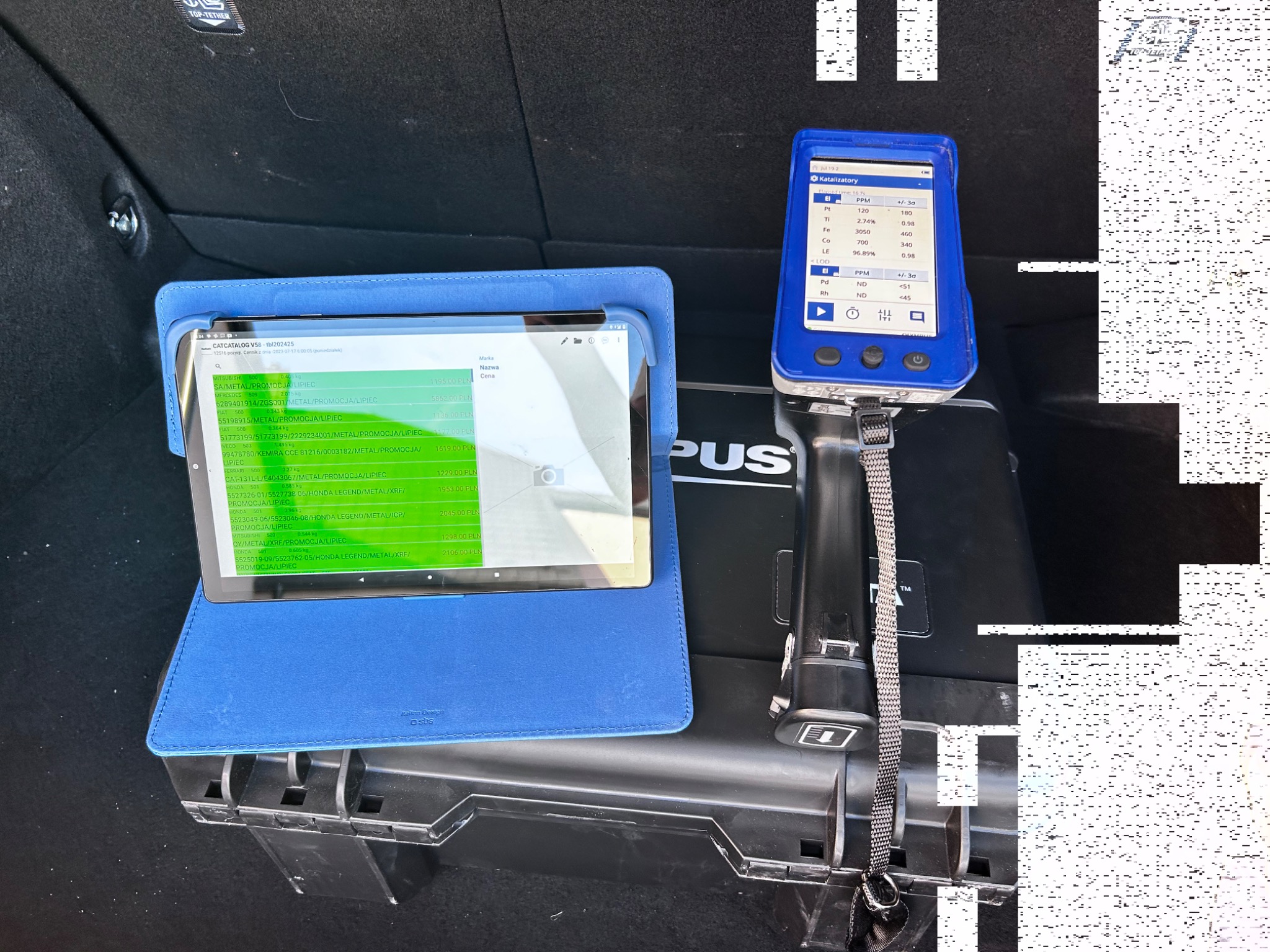 Tablet z otwartą aplikacją prezentującą listę katalizatorów z cenami, obok analizator metali z wynikami pomiarów w bagażniku samochodu.
