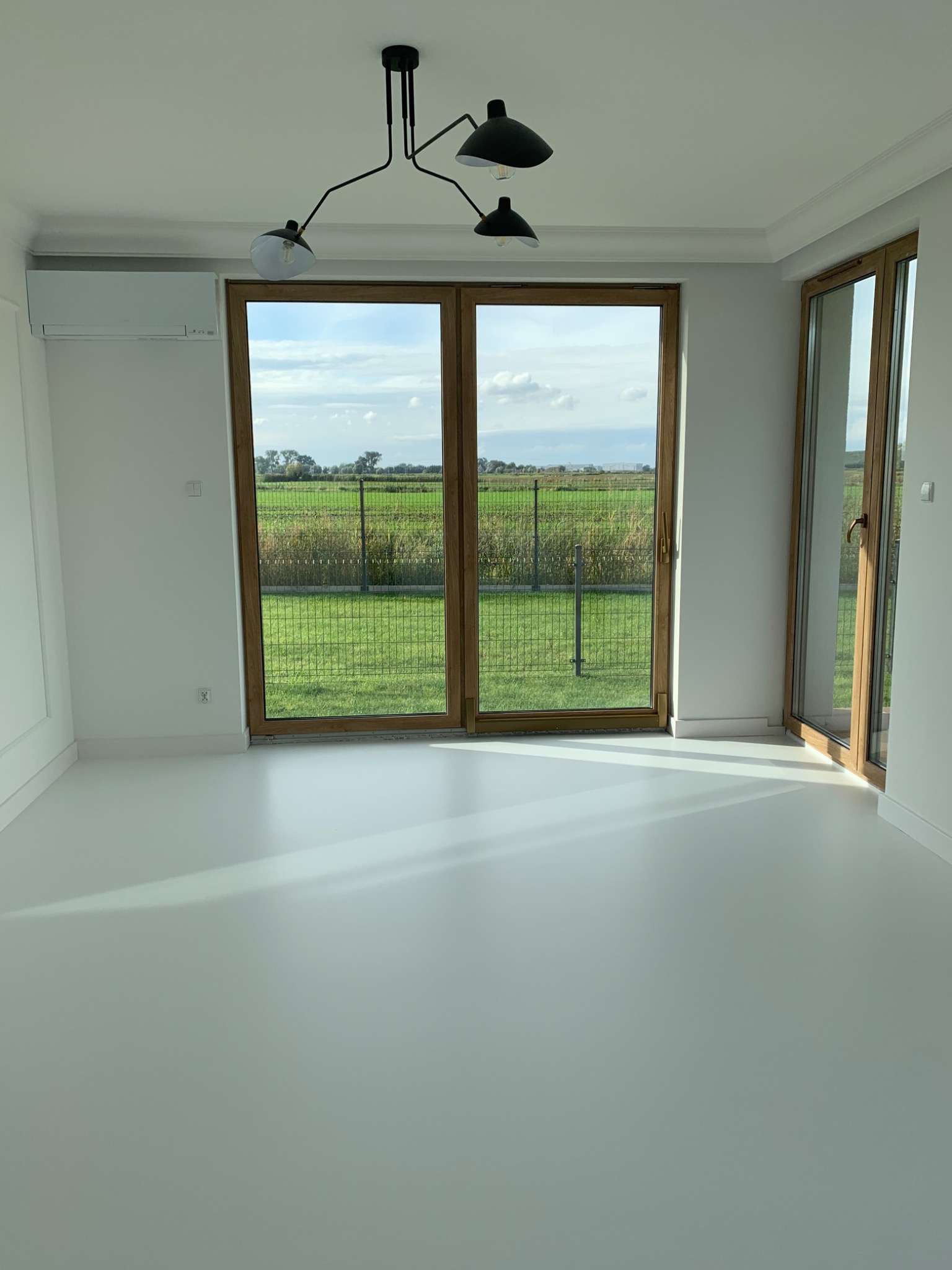 Jasne, puste pomieszczenie z białą, błyszczącą podłogą, widok na zewnątrz przez duże okna z drewnianymi ramami, nowoczesna lampa sufitowa.