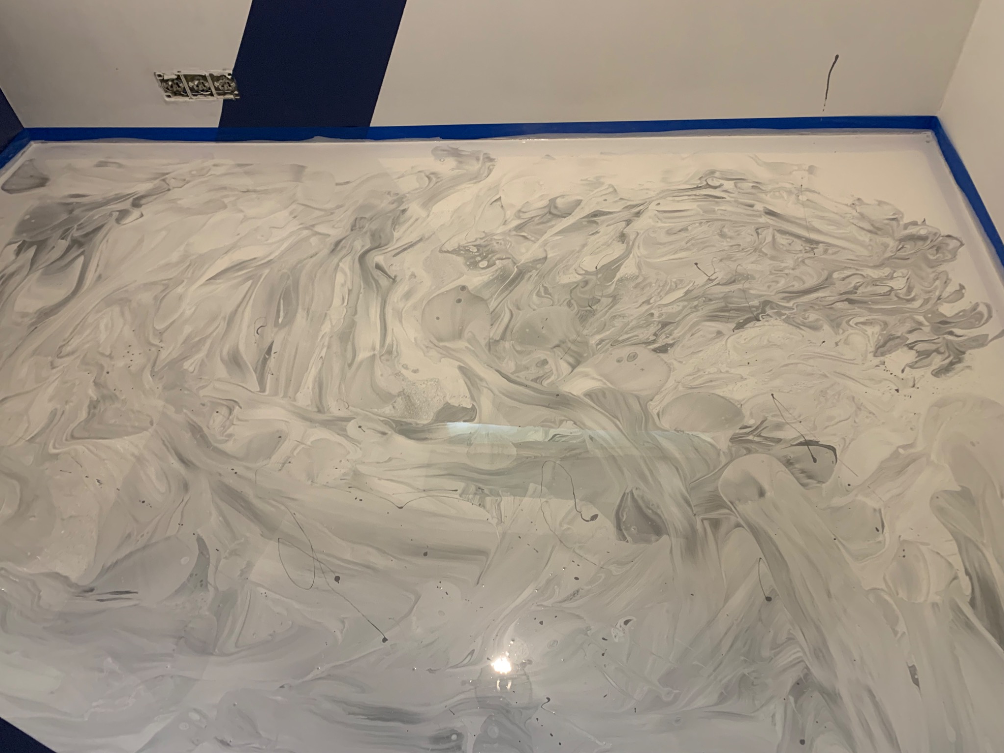 Świeżo wylana, wzorzysta podłoga w odcieniach szarości i bieli, zabezpieczona niebieską taśmą malarską przy ścianach, widok z góry, gniazdko elektryczne wystaje ze ściany, Wejherowo.