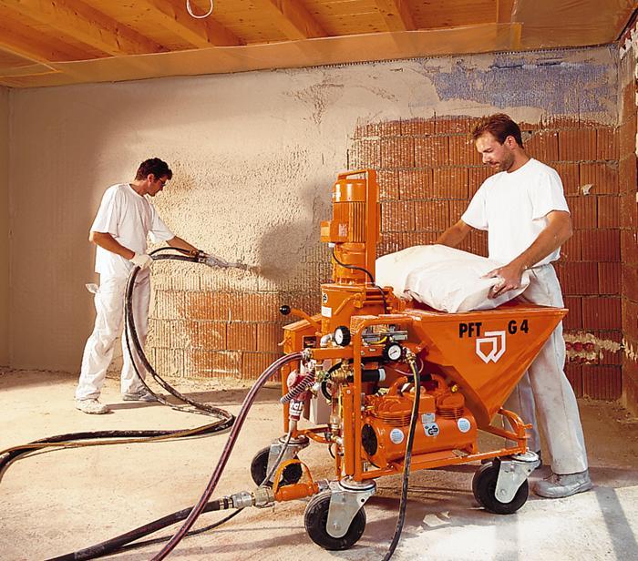 Dwóch pracowników w białych kombinezonach aplikuje tynk maszynowy na ceglaną ścianę za pomocą pomarańczowego agregatu PFT G4, widoczne węże i worki z zaprawą.