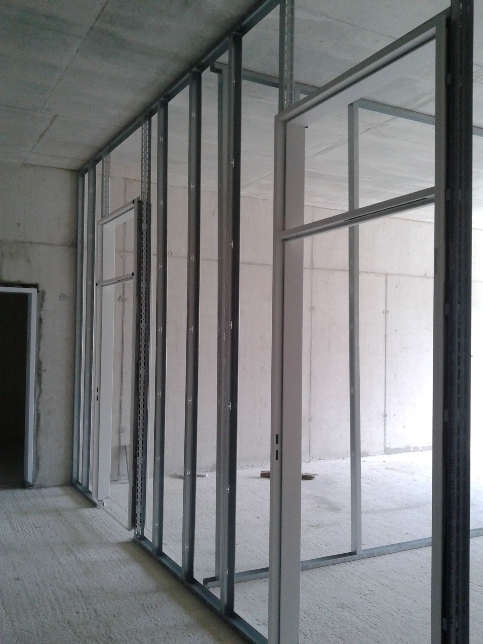 Metalowa konstrukcja szkieletowa ścian działowych z otworami na drzwi, widoczna w surowym stanie, na tle betonowej podłogi i ścian w trakcie budowy.