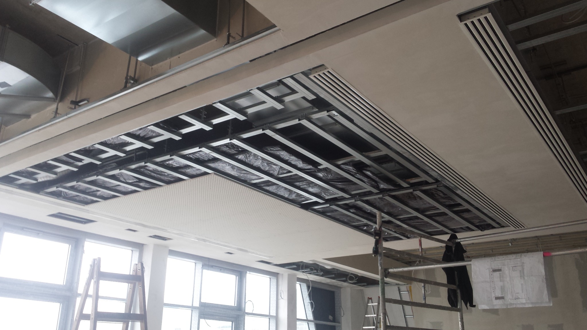 Montaż sufitu podwieszanego z widoczną konstrukcją metalową, izolacją i częściowo zamontowanymi panelami akustycznymi w przestrzeni biurowej, z użyciem rusztowania i planów technicznych.