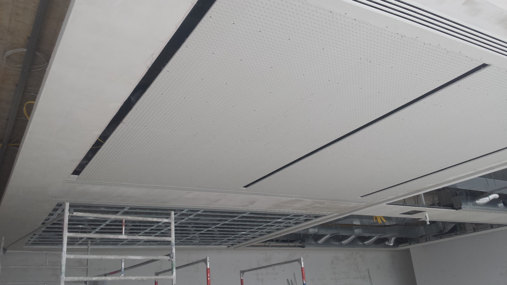 Montaż sufitu podwieszanego z płyt perforowanych z widoczną konstrukcją nośną i elementami wentylacji, częściowo zakryty rusztowaniem.