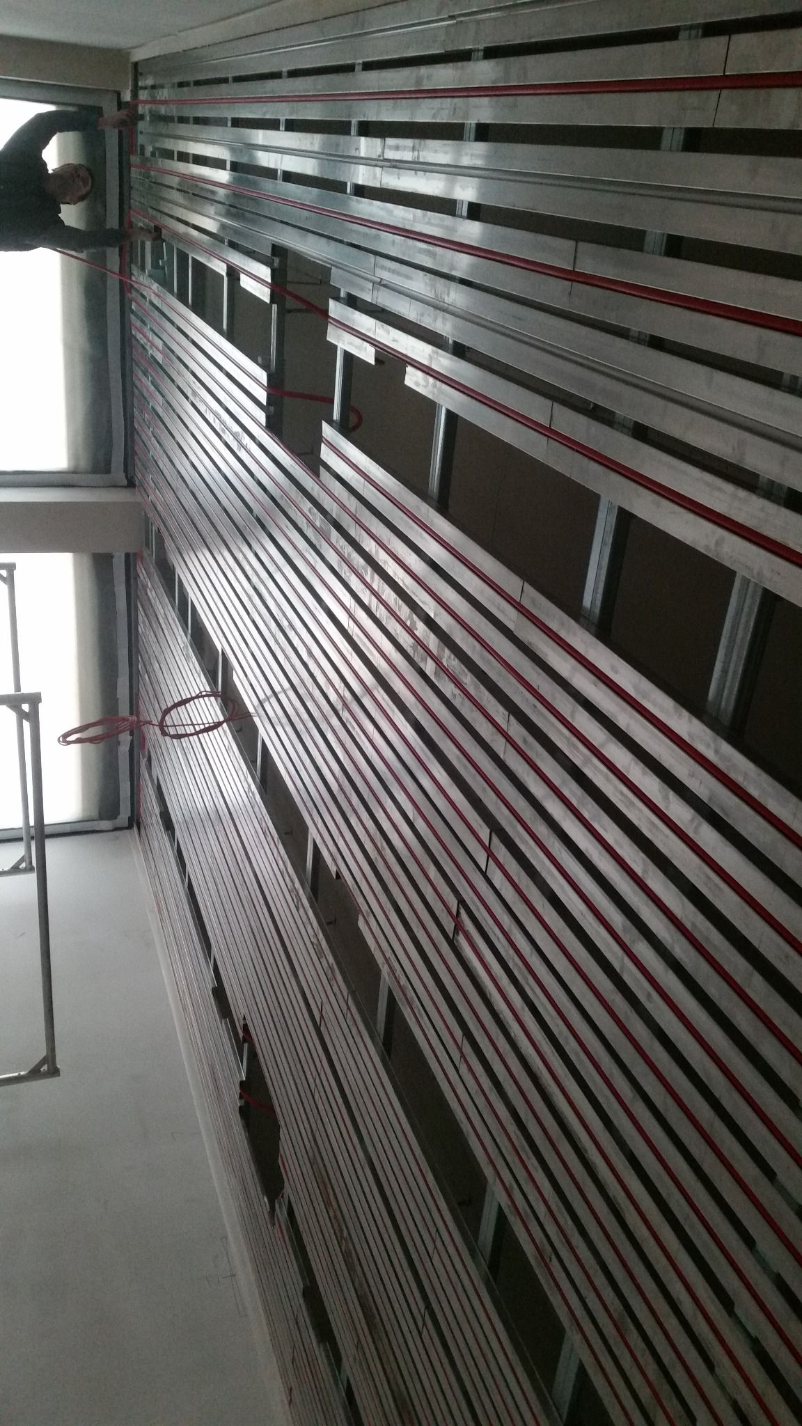 Montaż stelażu z profili metalowych z czerwonymi przewodami wzdłuż ściany, widoczne okno i fragment rusztowania.