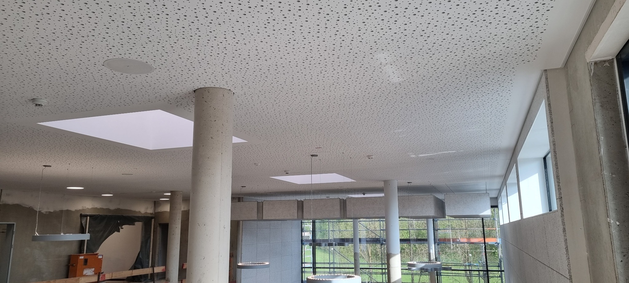 Montaż perforowanego sufitu podwieszanego z okrągłymi lampami wiszącymi i betonowymi kolumnami w przestronnym wnętrzu w trakcie wykończenia.