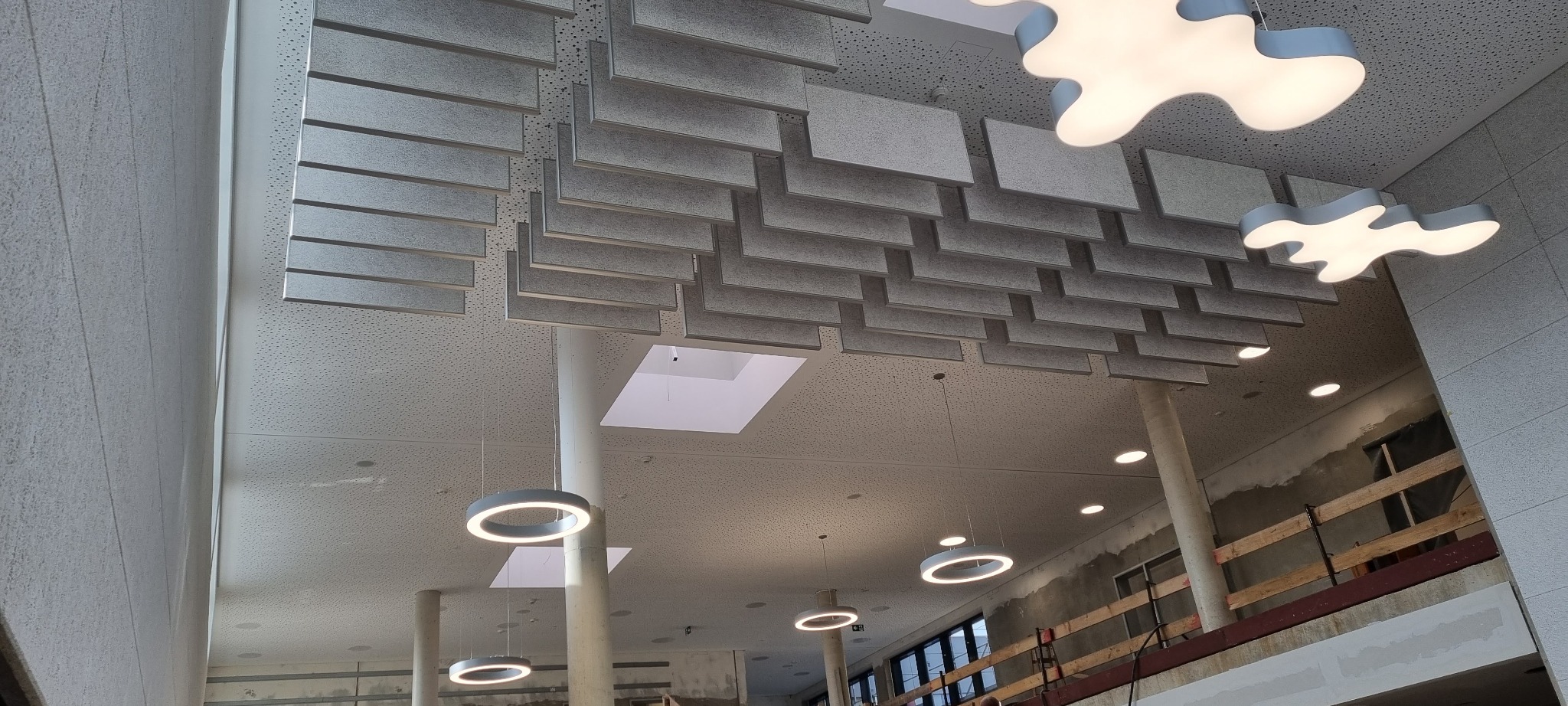 Sufit podwieszany z paneli akustycznych w biurze, oświetlenie LED w kształcie chmur, widoczne elementy konstrukcyjne w tle.