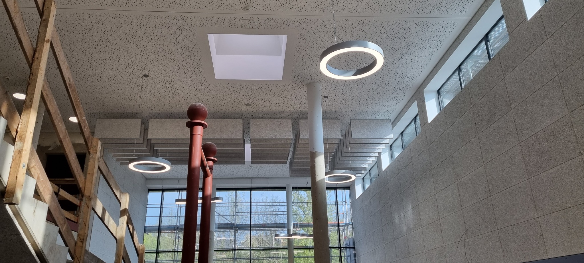 Wnętrze budynku w Proszowicach z elementami wykończenia: podwieszany sufit z panelami akustycznymi, okrągłe lampy LED, ściana wyłożona panelami oraz drewniana konstrukcja schodów w trakcie montażu.