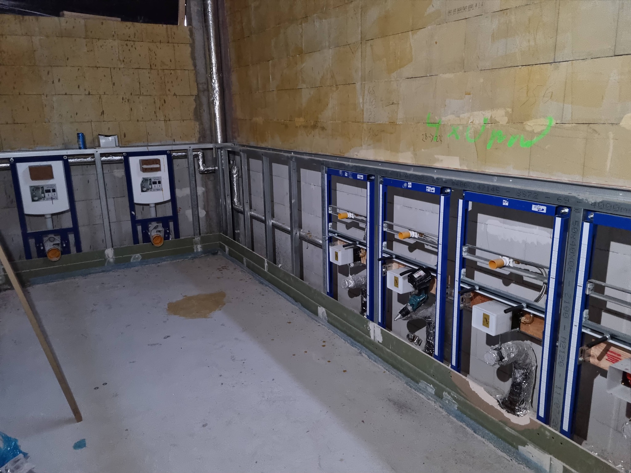 Wnętrze łazienki w trakcie remontu: widoczne stelaże podtynkowe WC zamontowane na ścianie z bloczków, z widocznymi instalacjami hydraulicznymi i wentylacyjnymi, betonowa wylewka podłogowa...