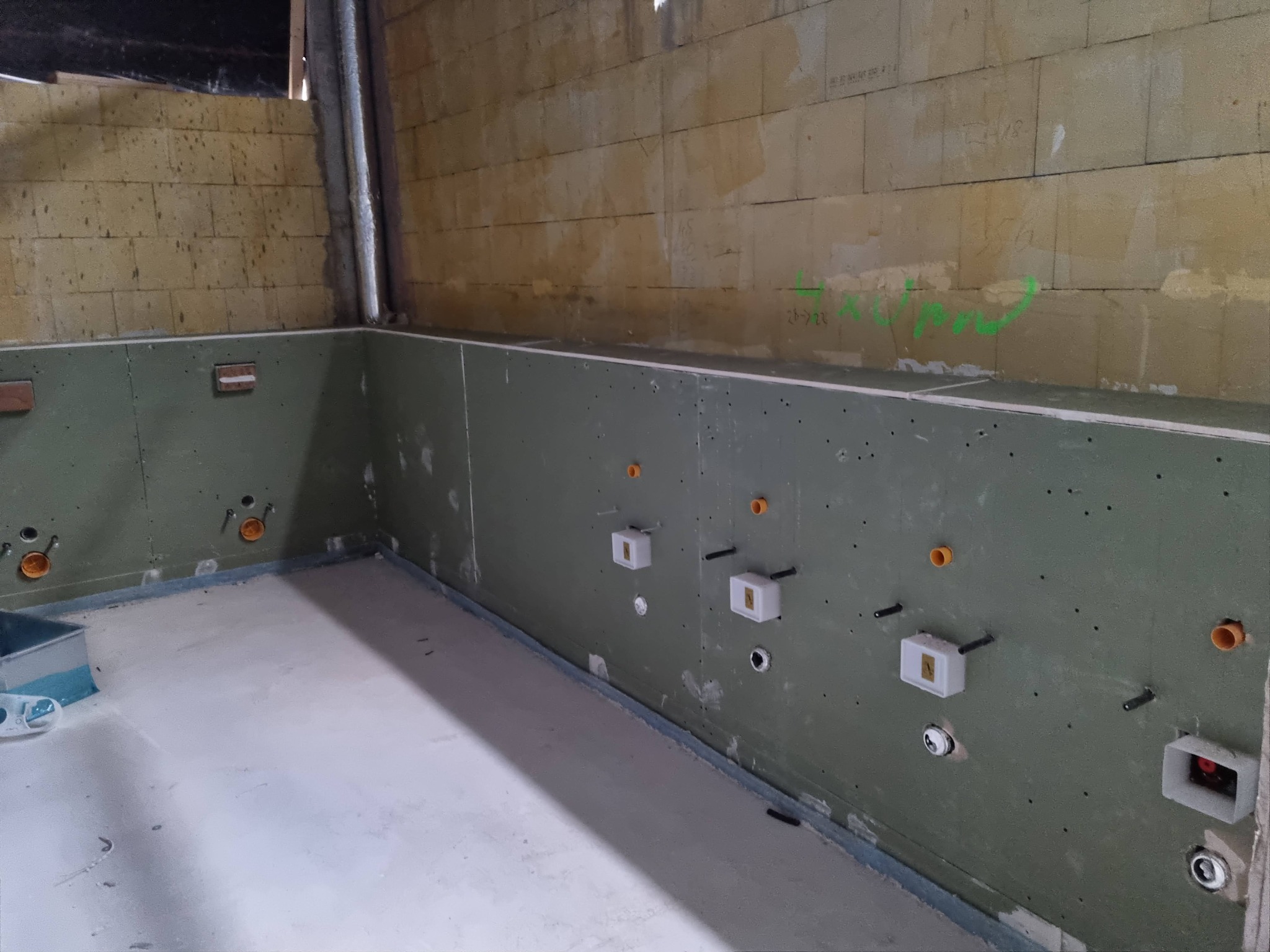 Wnętrze w trakcie prac wykończeniowych: zielona płyta na ścianach z widocznymi instalacjami hydraulicznymi i elektrycznymi, surowy mur z bloków betonowych powyżej, wylewka podłogowa.