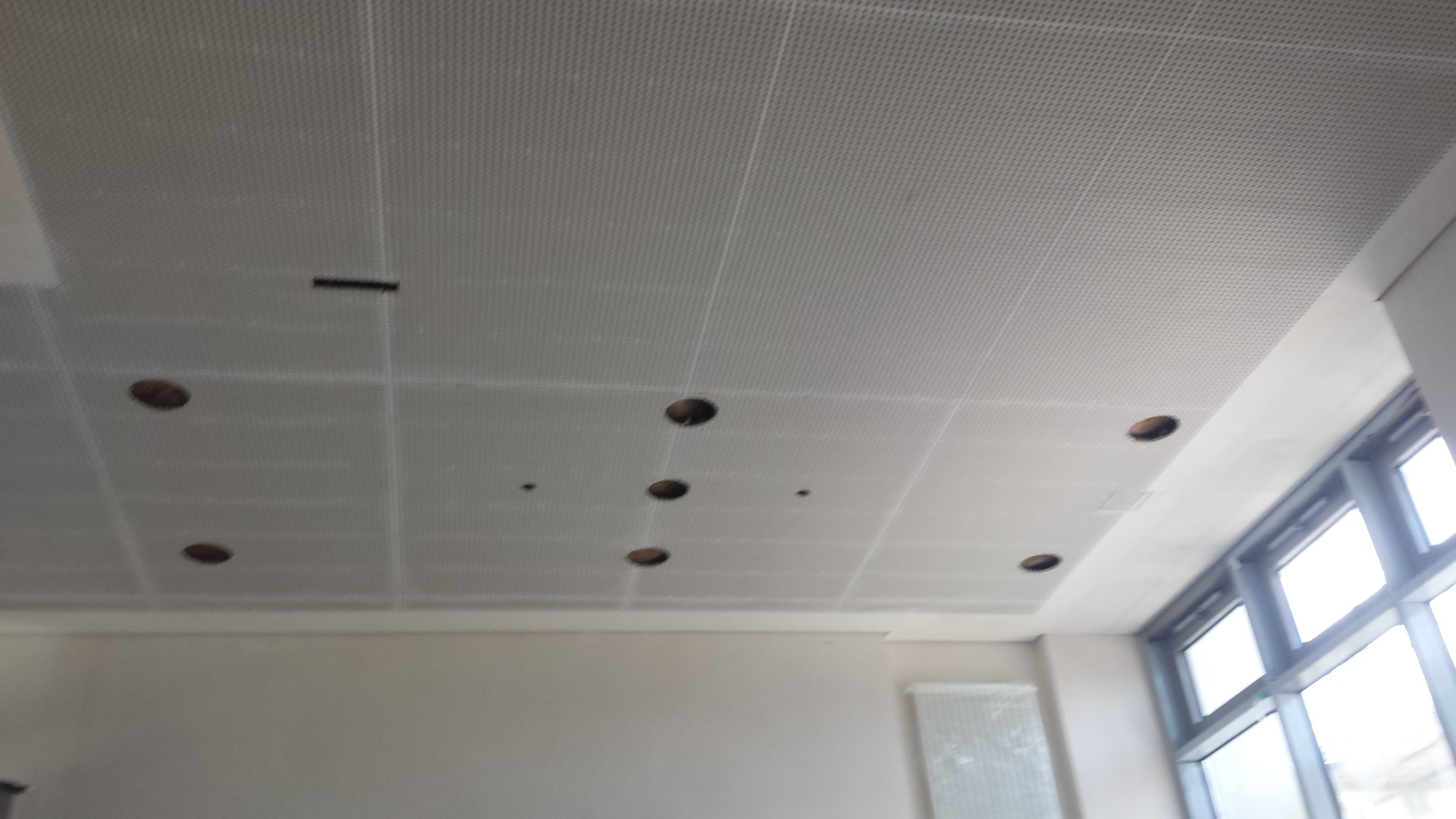 Biały sufit podwieszany z perforowanej płyty z widocznymi otworami na oświetlenie, zarys okna w tle po prawej stronie.