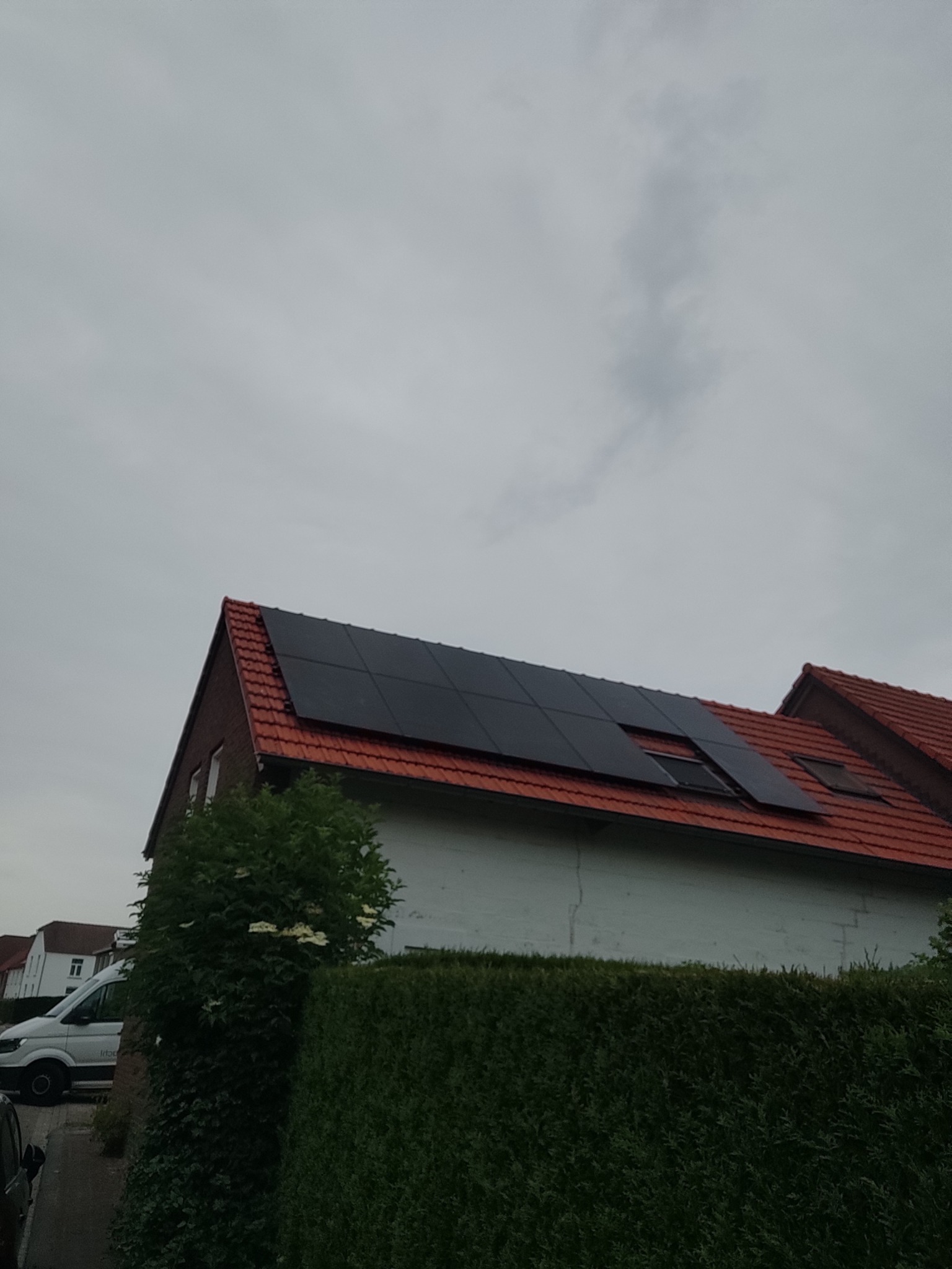 Ciemne panele na dachu z czerwonej dachówki, częściowo zasłonięte przez krzewy, na tle pochmurnego nieba.