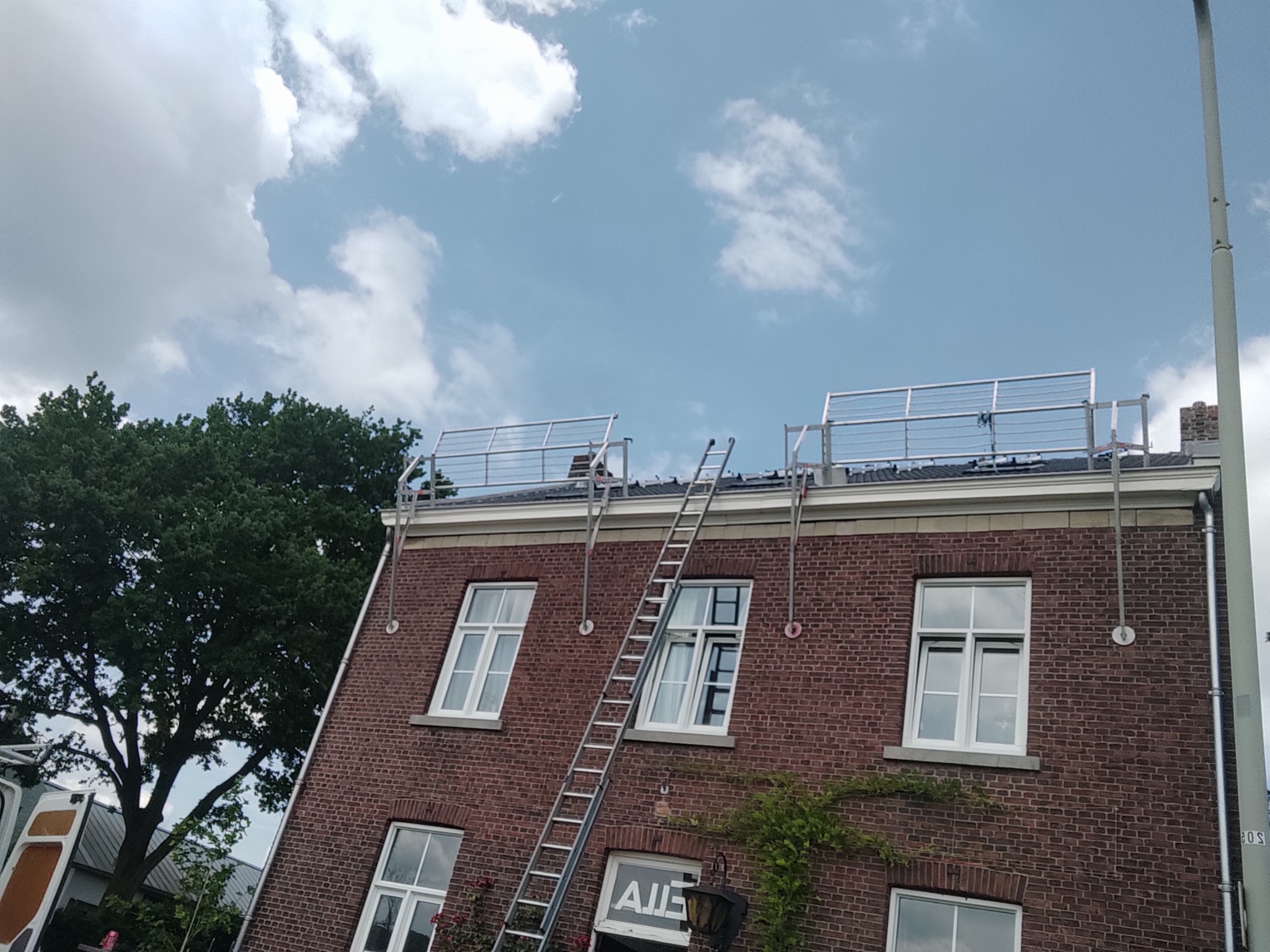 Montaż konstrukcji wsporczej na dachu ceglanego budynku, widoczna drabina oparta o ścianę, błękitne niebo z chmurami w tle.