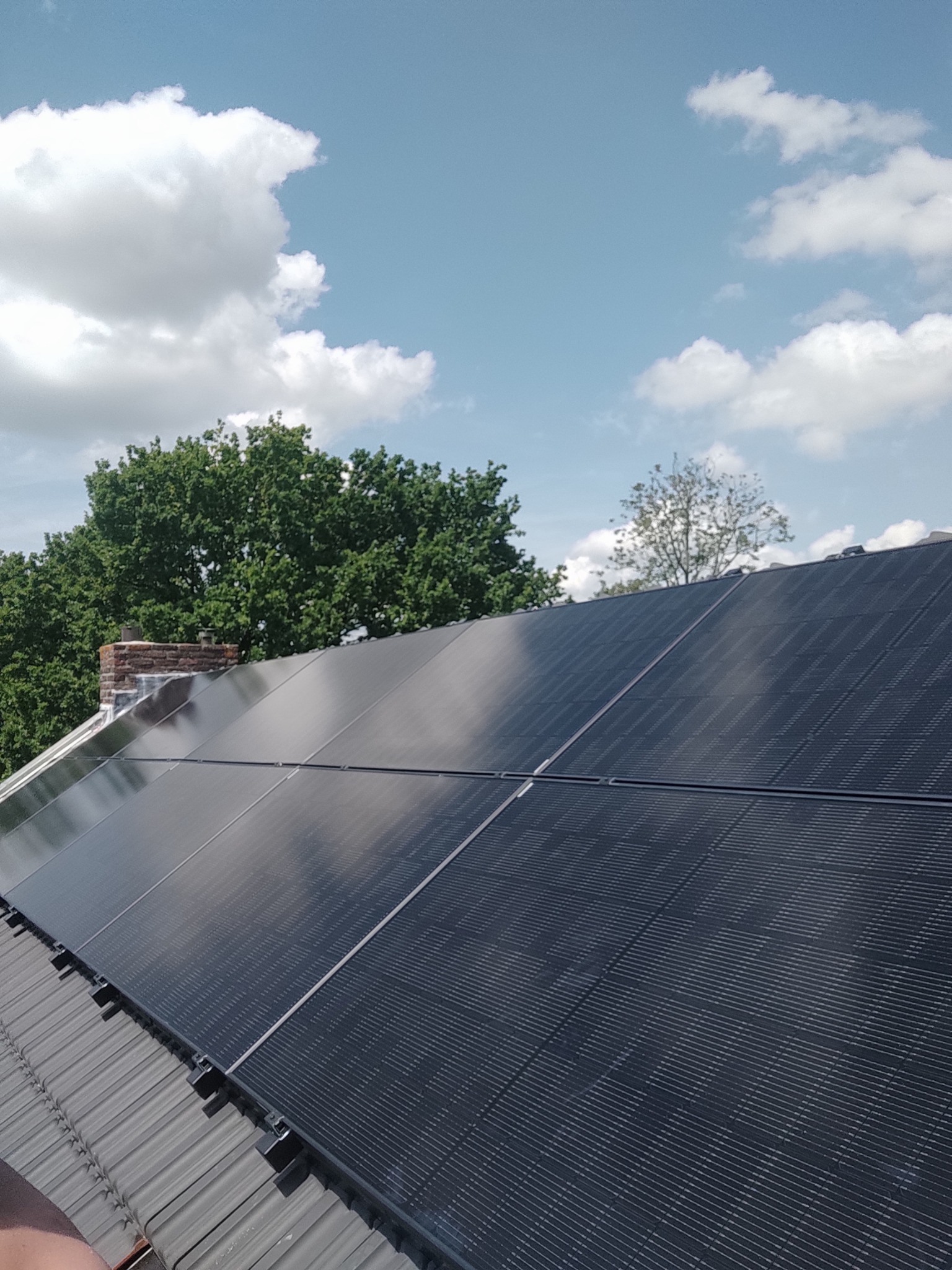 Panele słoneczne na dachu domu w Brunssum, widok z dołu pod kątem, z kominem i drzewami w tle oraz fragmentem błękitnego nieba z chmurami.