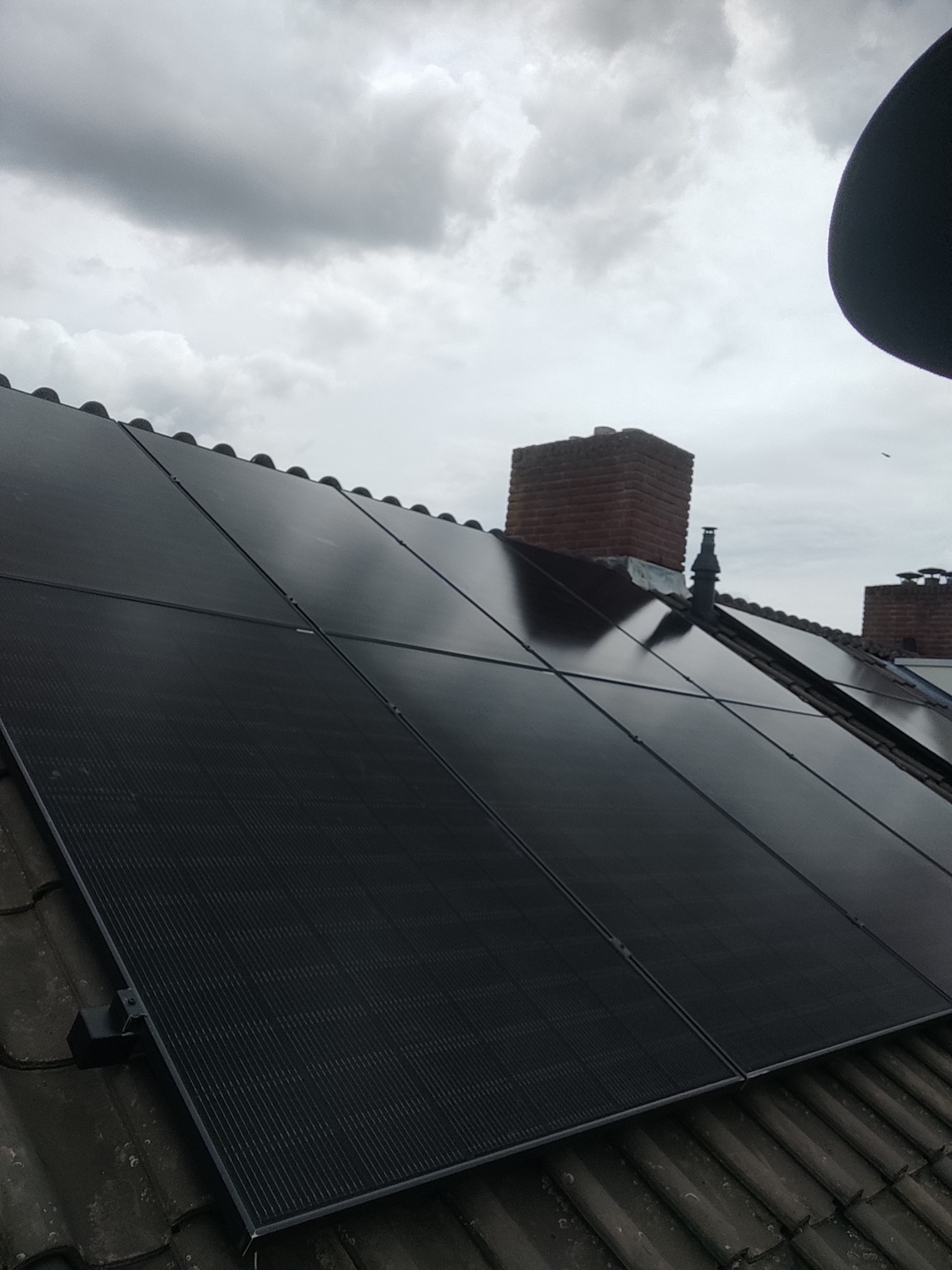 Ciemne panele słoneczne na dachu z dachówki ceramicznej, widoczne kominy z cegły na tle pochmurnego nieba. Zastosowano czarne mocowania paneli.